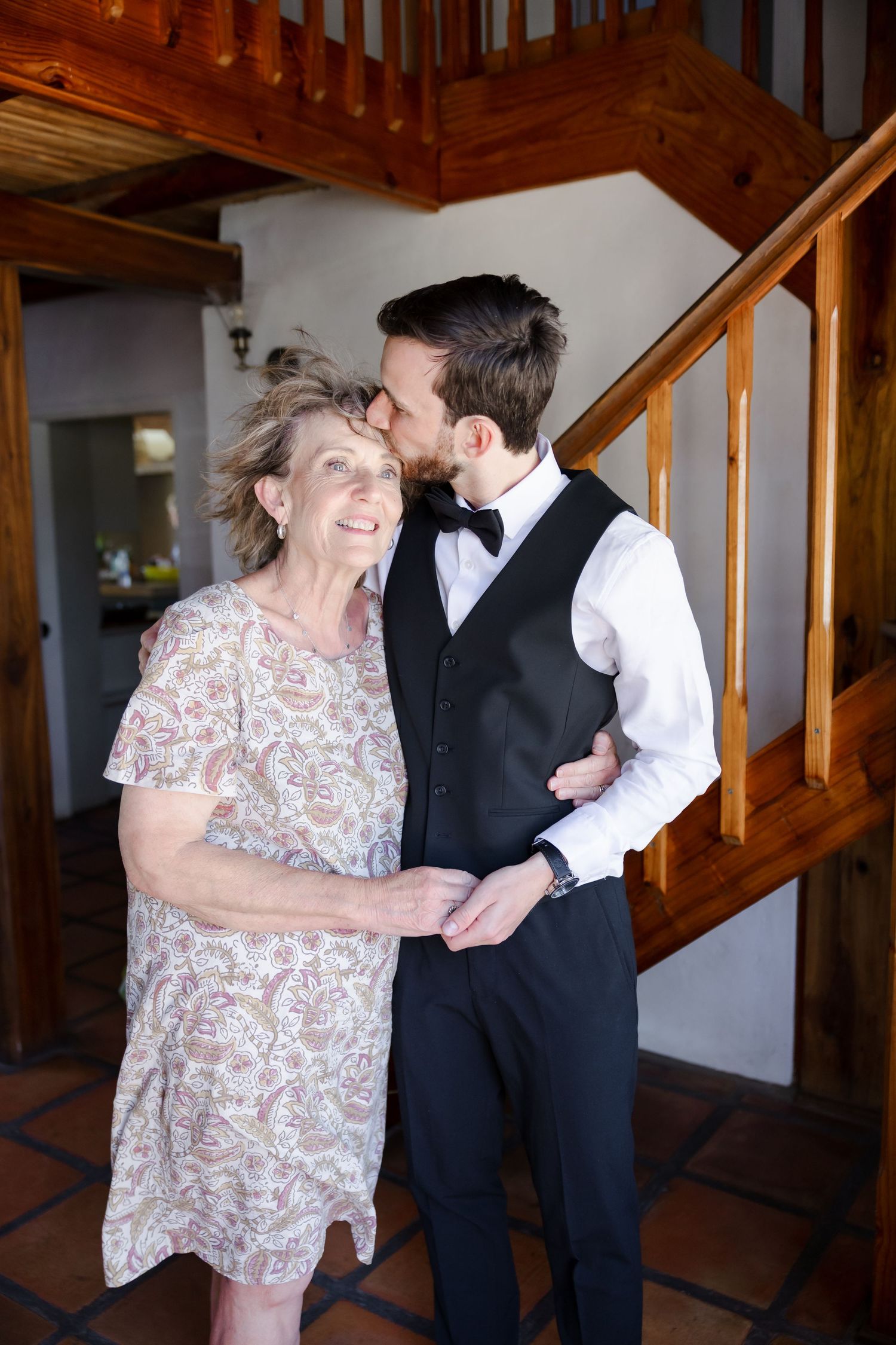 A tender moment shared between two people in formal attire near a wooden staircase in an indoor setting.