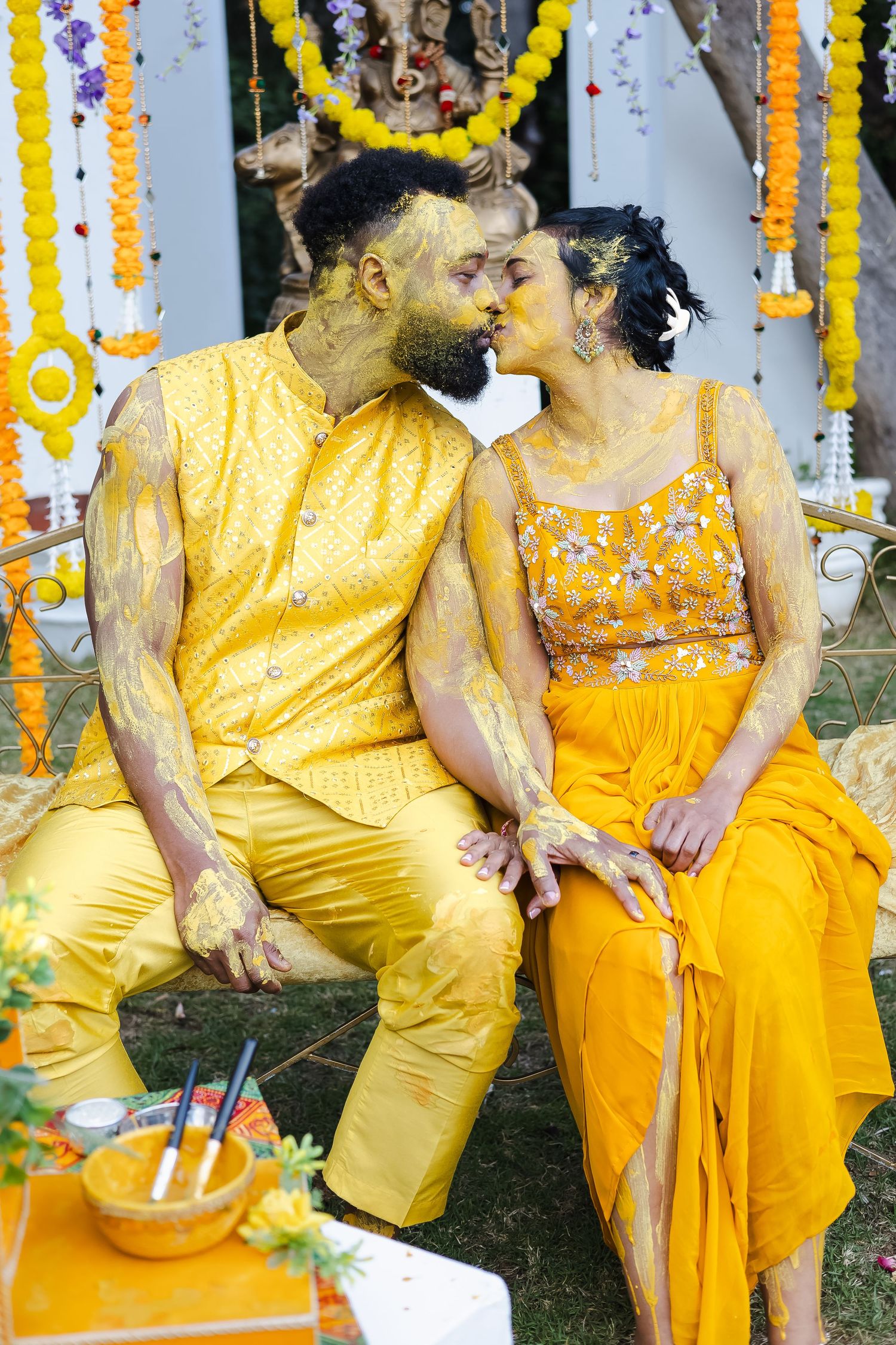 Wedding guests in coordinated yellow and orange outfits sit together during an outdoor celebration with floral decorations.