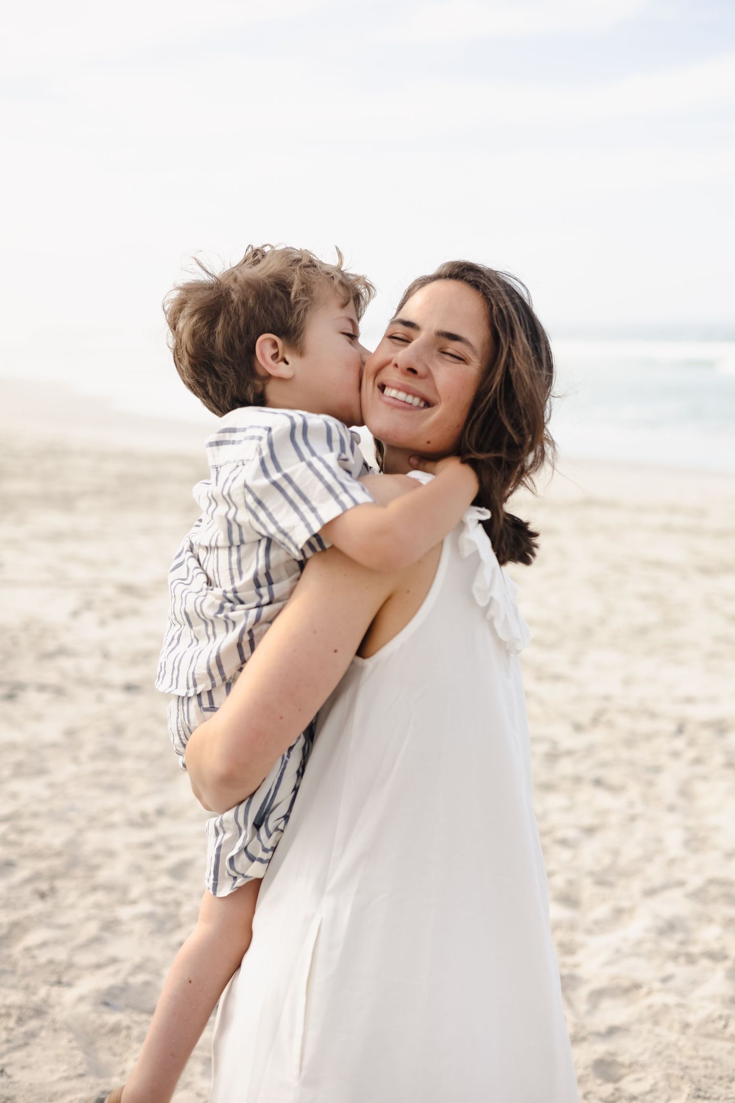 A young mother in a white dress holds her son on a sandy beach as he gives her a sweet kiss on the cheek.
