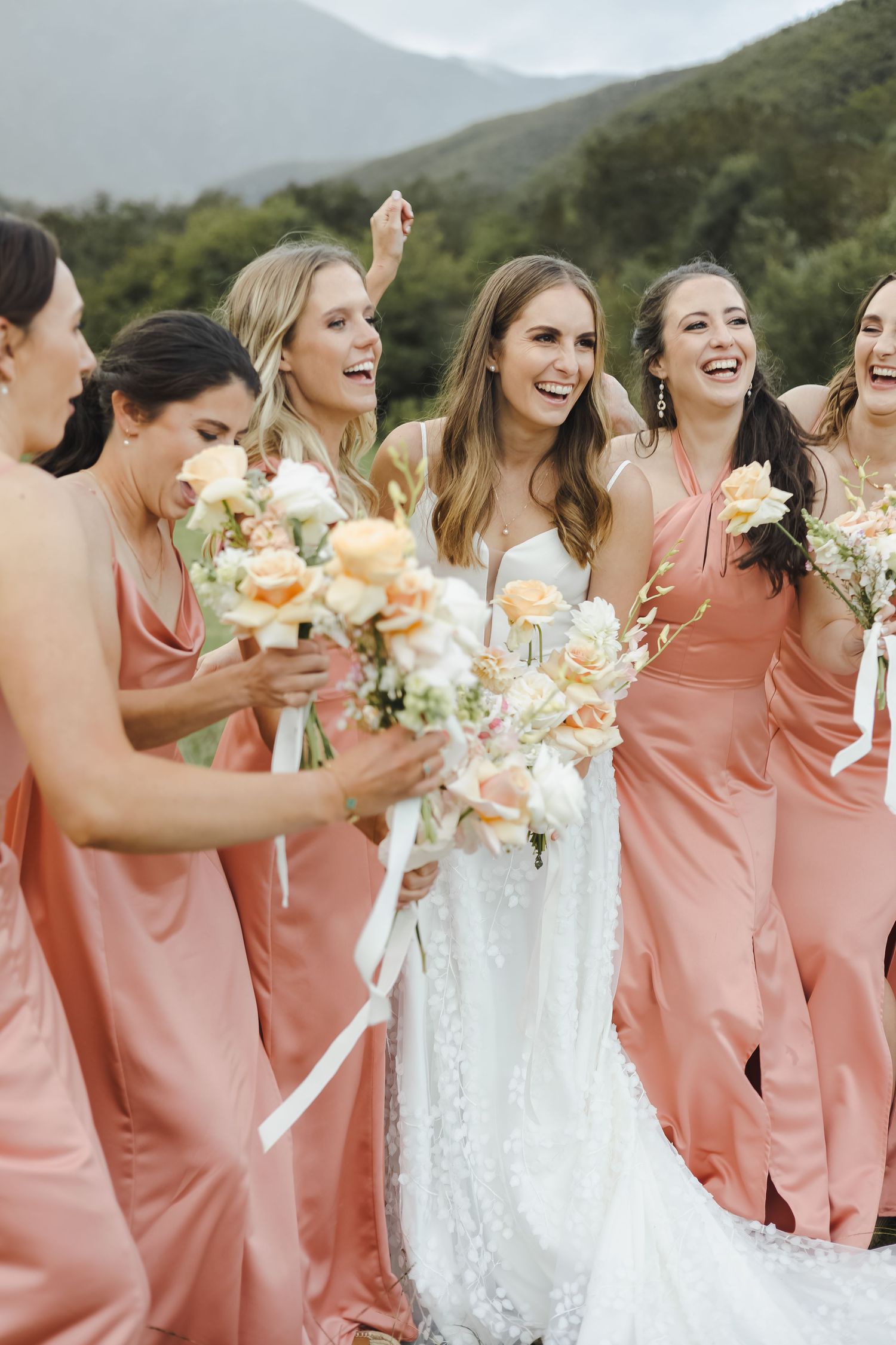Joyful outdoor wedding celebration with attendants in coral dresses surrounded by mountains.