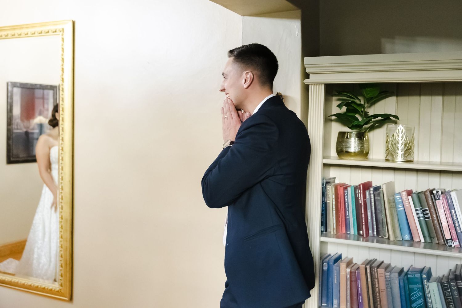 Person in a navy suit stands in a home library or office space with bookshelves and framed artwork visible.
