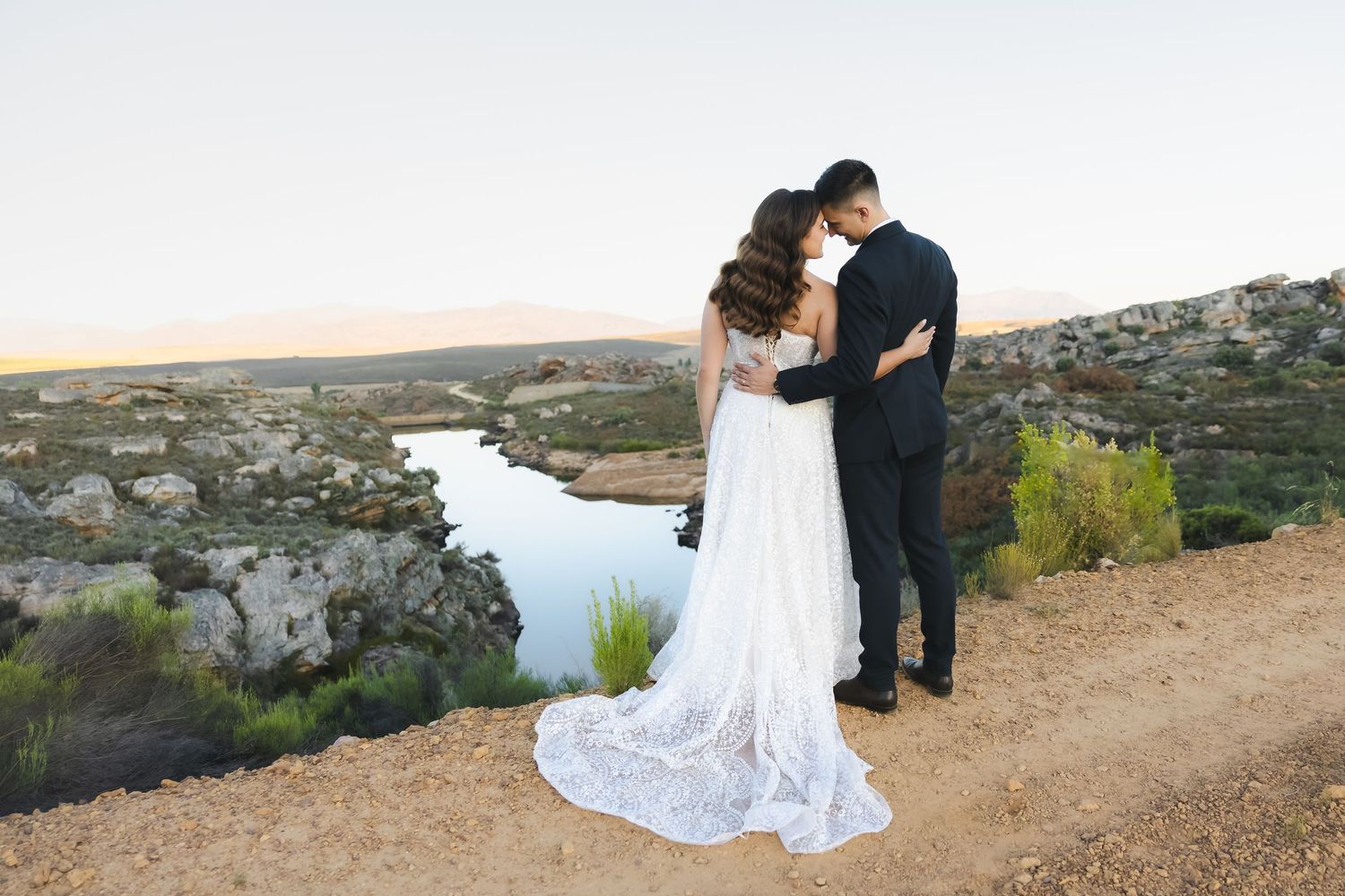 Wedding couple shares intimate moment overlooking scenic river valley with rocky cliffs at sunset.