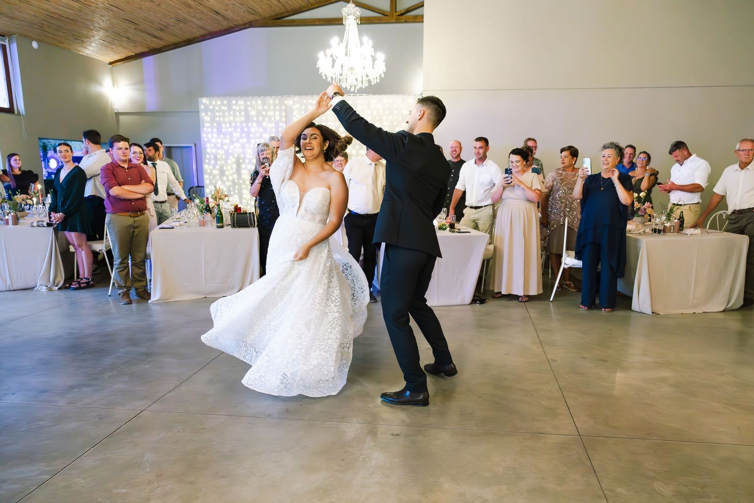 A couple performs their first dance at a wedding reception in a rustic venue with wood paneled ceiling.