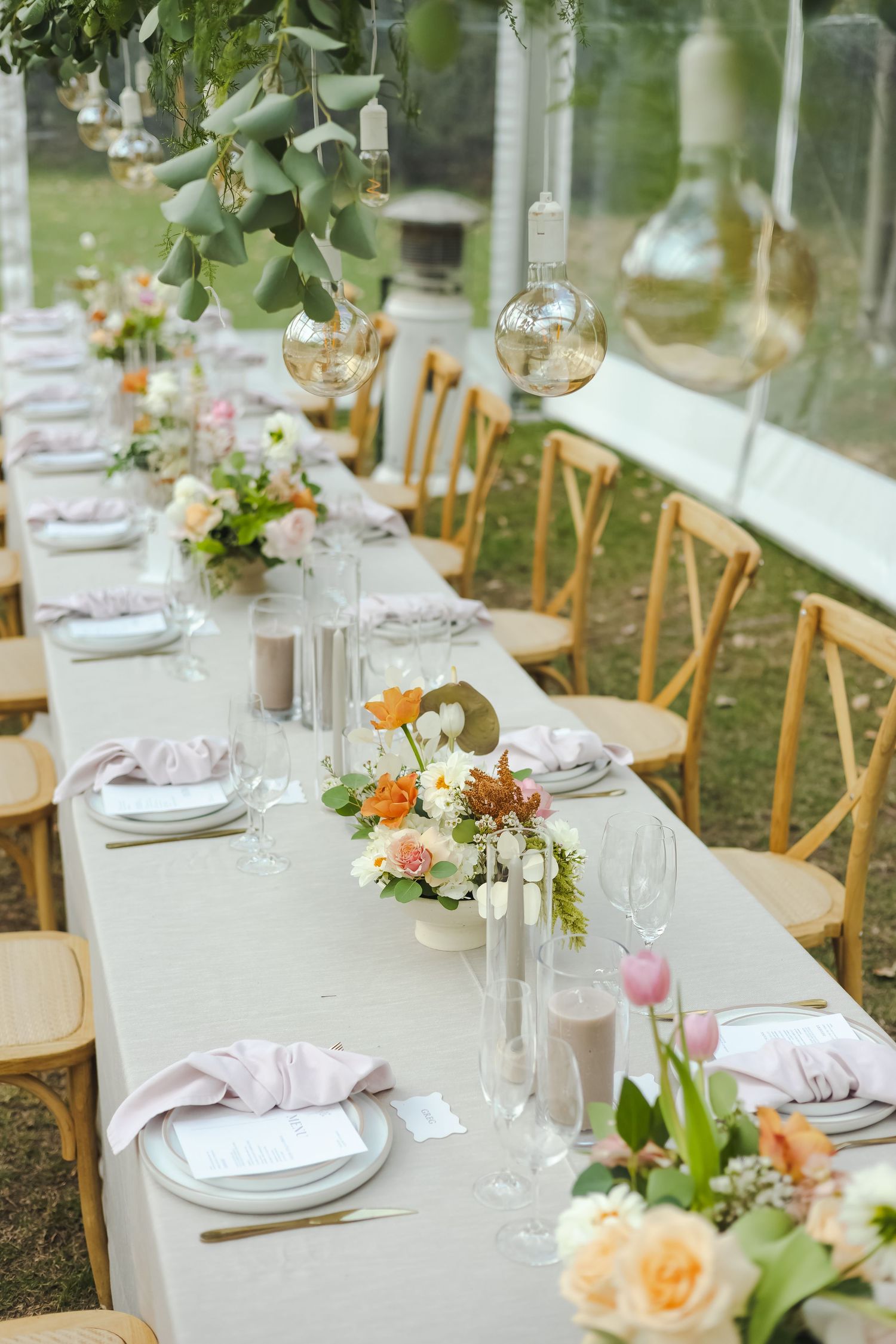 Long banquet tables set with white linens and floral centerpieces in a bright garden tent setting.