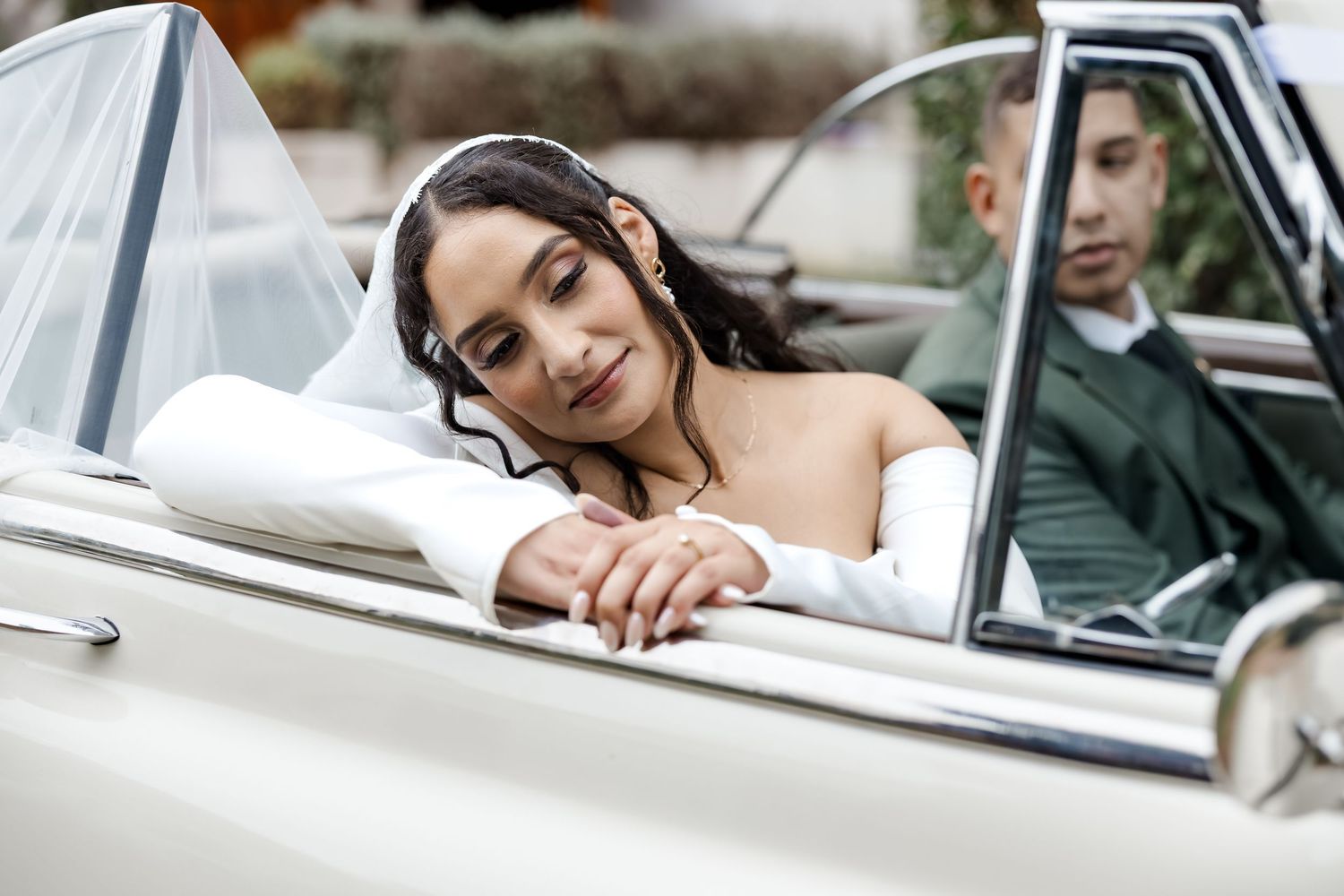 Bride in white dress leans on vintage convertible car door beside groom in green suit at wedding.