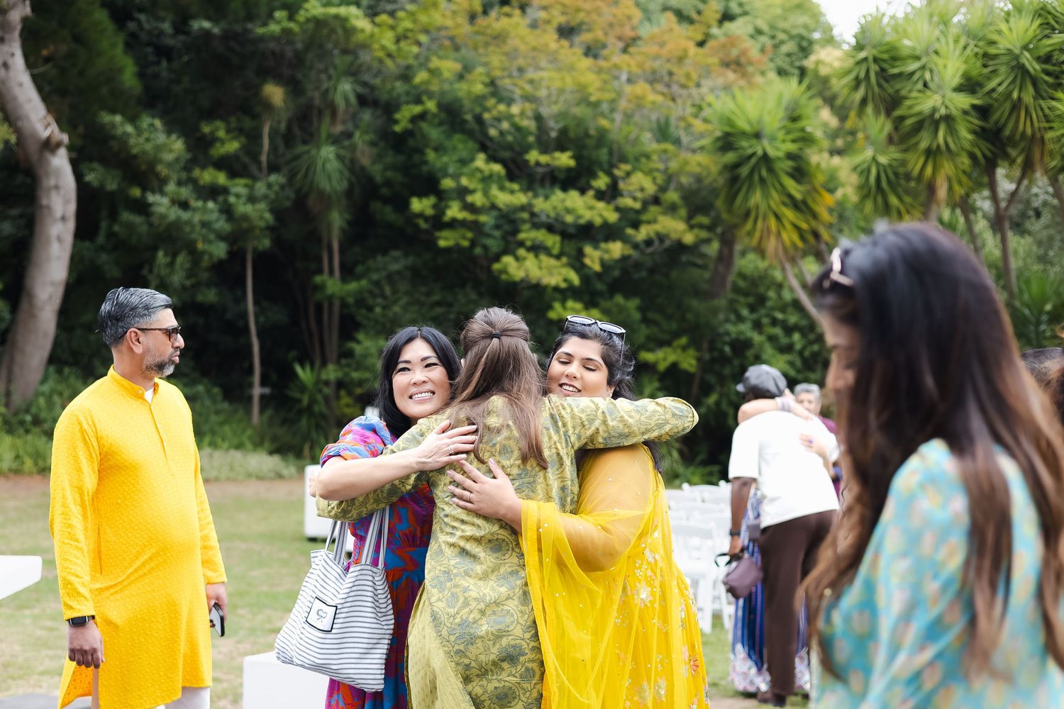 A group celebrates outdoors with hugs and bright yellow traditional attire among tropical greenery.