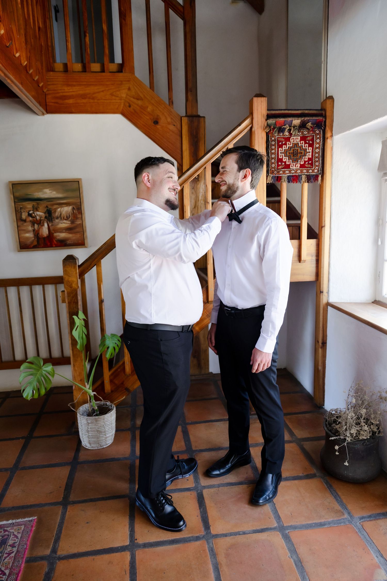 Two people in formal white shirts and black pants help each other get ready in an entryway with wooden stairs.