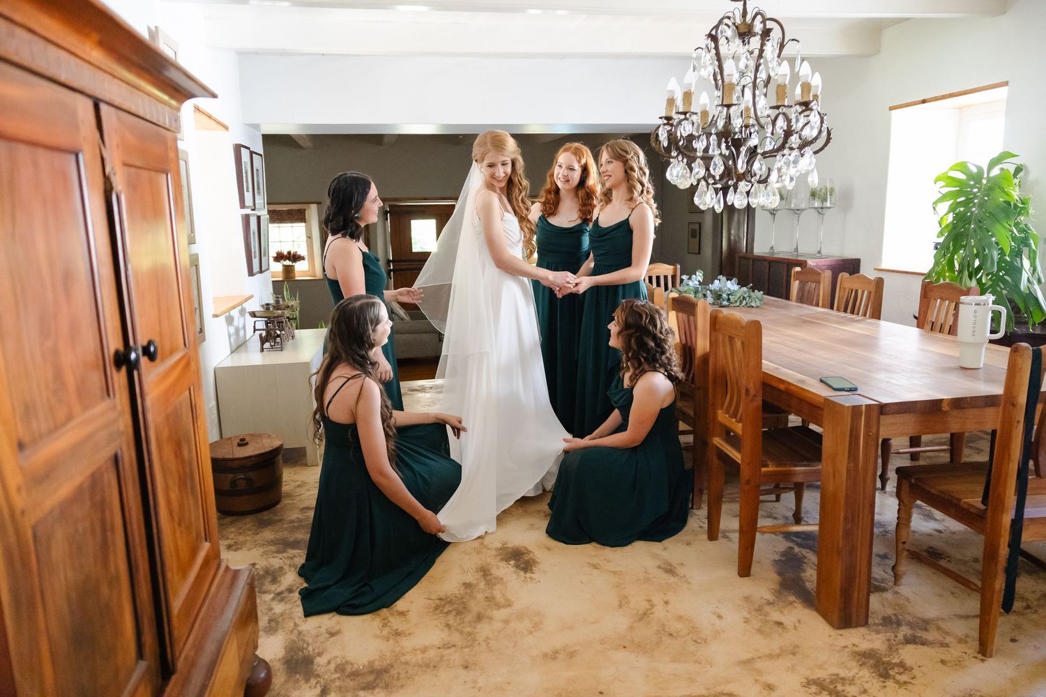 A bridal party in navy blue dresses helps adjust the bride's white gown in an elegant dining room with chandelier.
