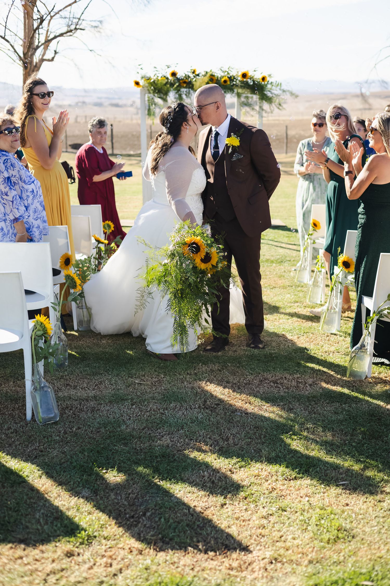 Bride and groom share first kiss at outdoor sunflower-themed wedding ceremony with guests applauding on sunny day.