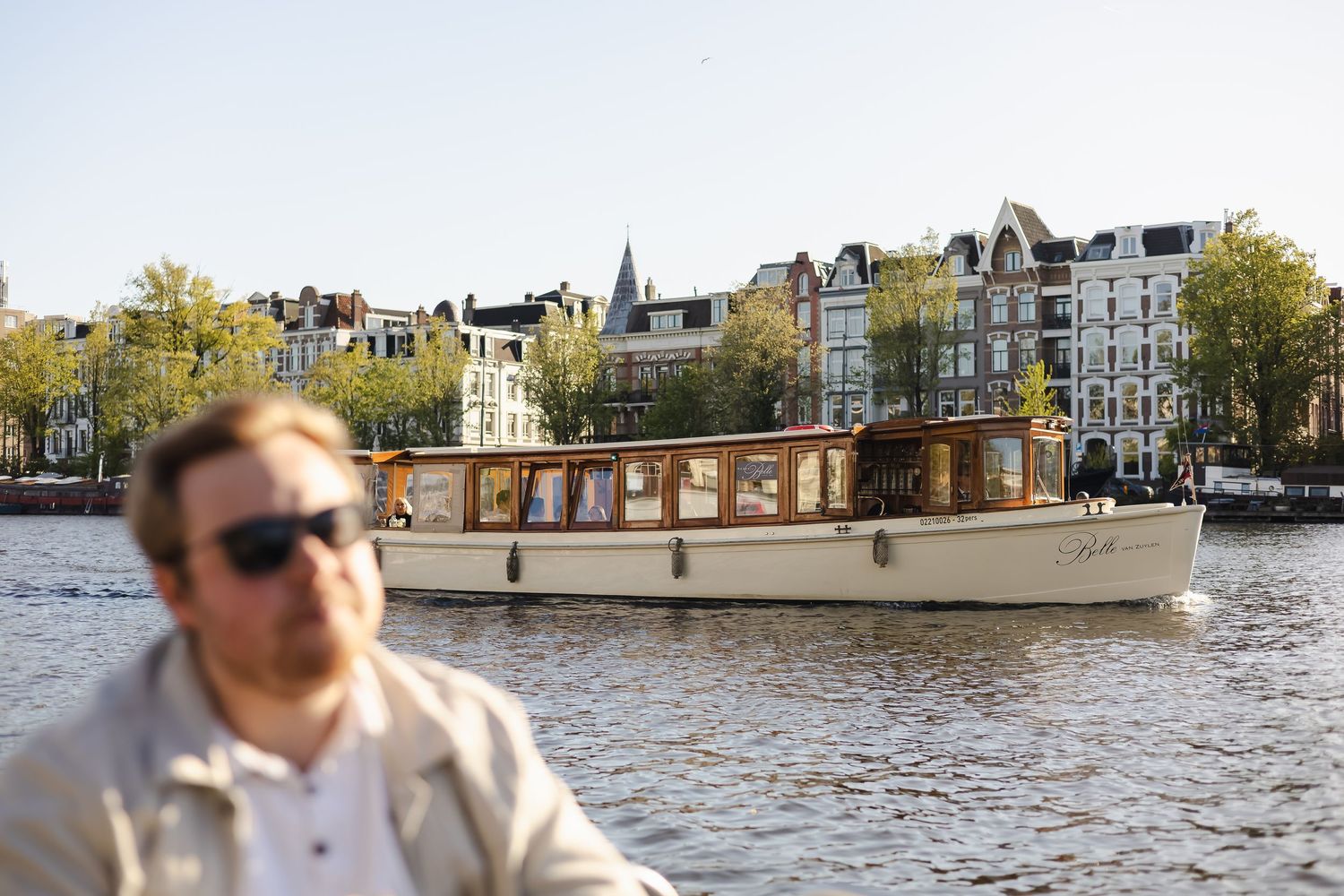 A man in sunglasses sits near a classic wooden canal boat cruising Amsterdam's waterways with historic buildings in background.