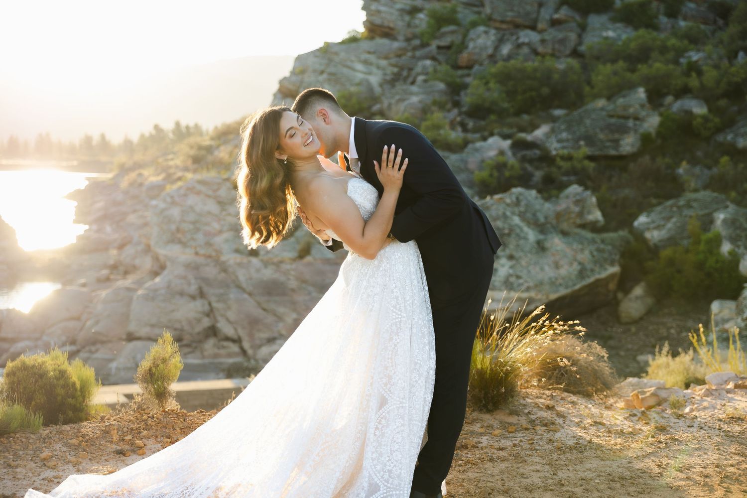 Couple in wedding attire embraces on rocky cliffside during golden sunset hour.