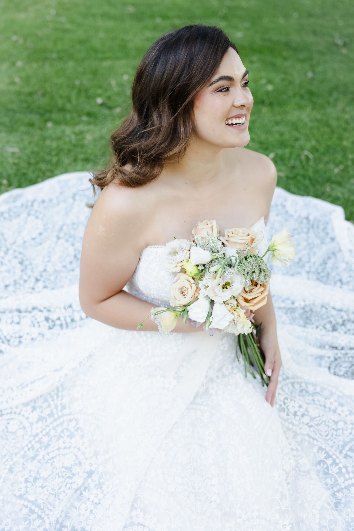 A bride in a white lace gown lies on grass holding a delicate bouquet of white and peach flowers during a wedding photoshoot.