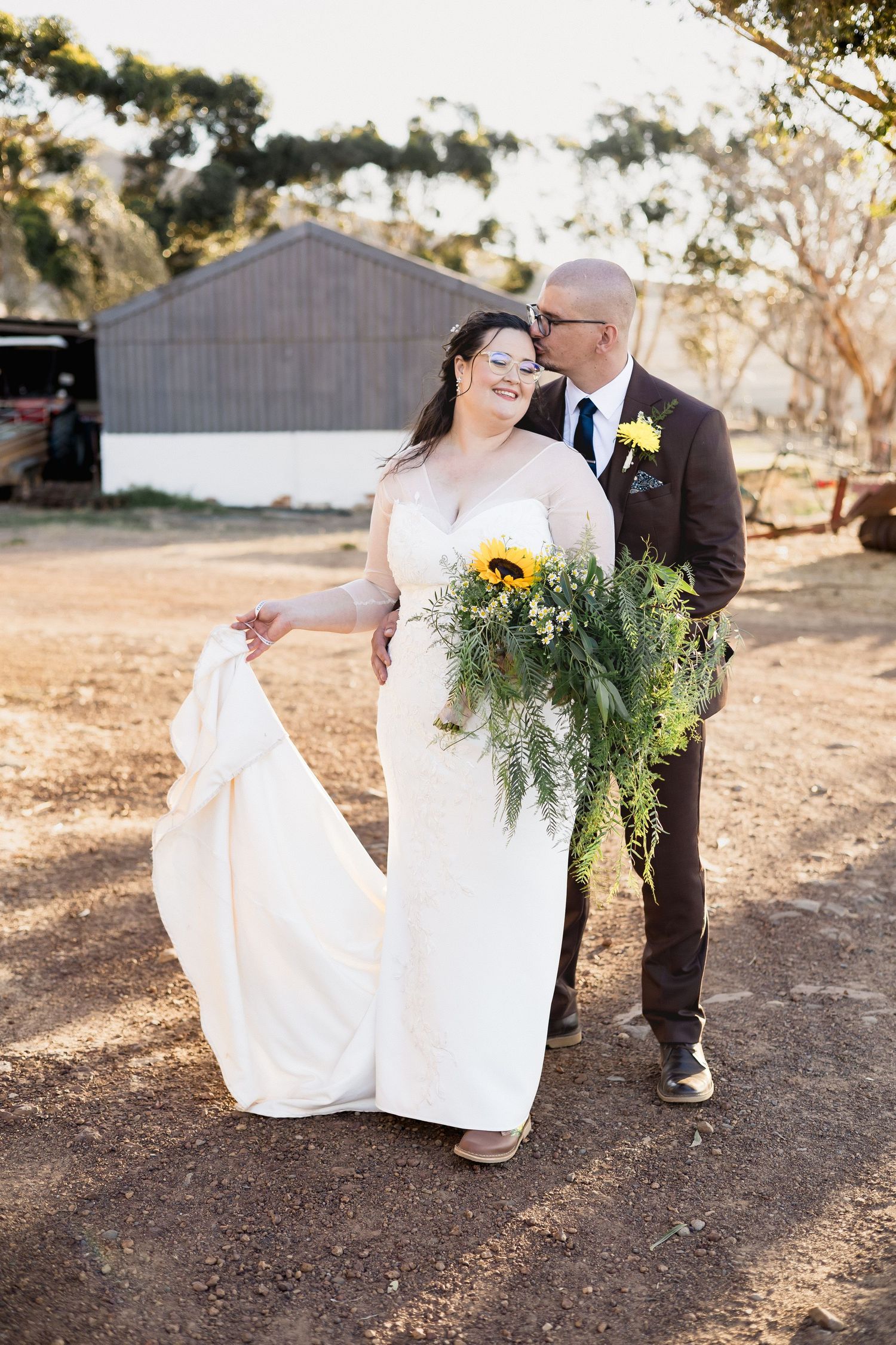 Bride and groom share a kiss outdoors on their wedding day, with sunflower bouquet and rustic barn backdrop.