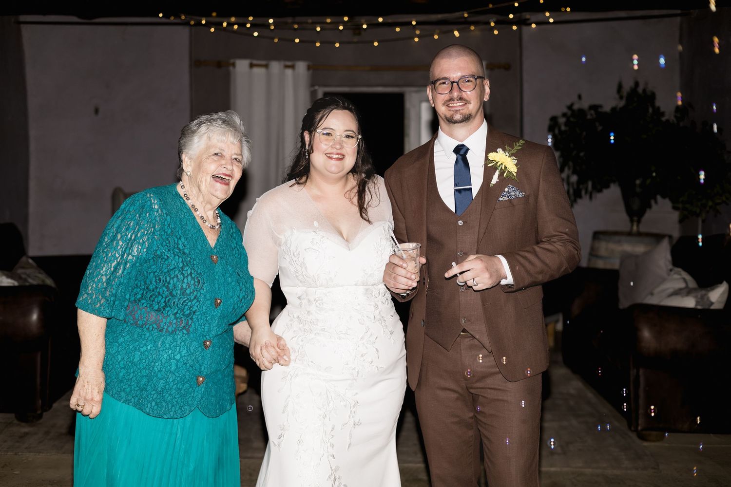 Bride in white gown poses with groom in brown suit and elderly woman in teal dress at wedding reception.