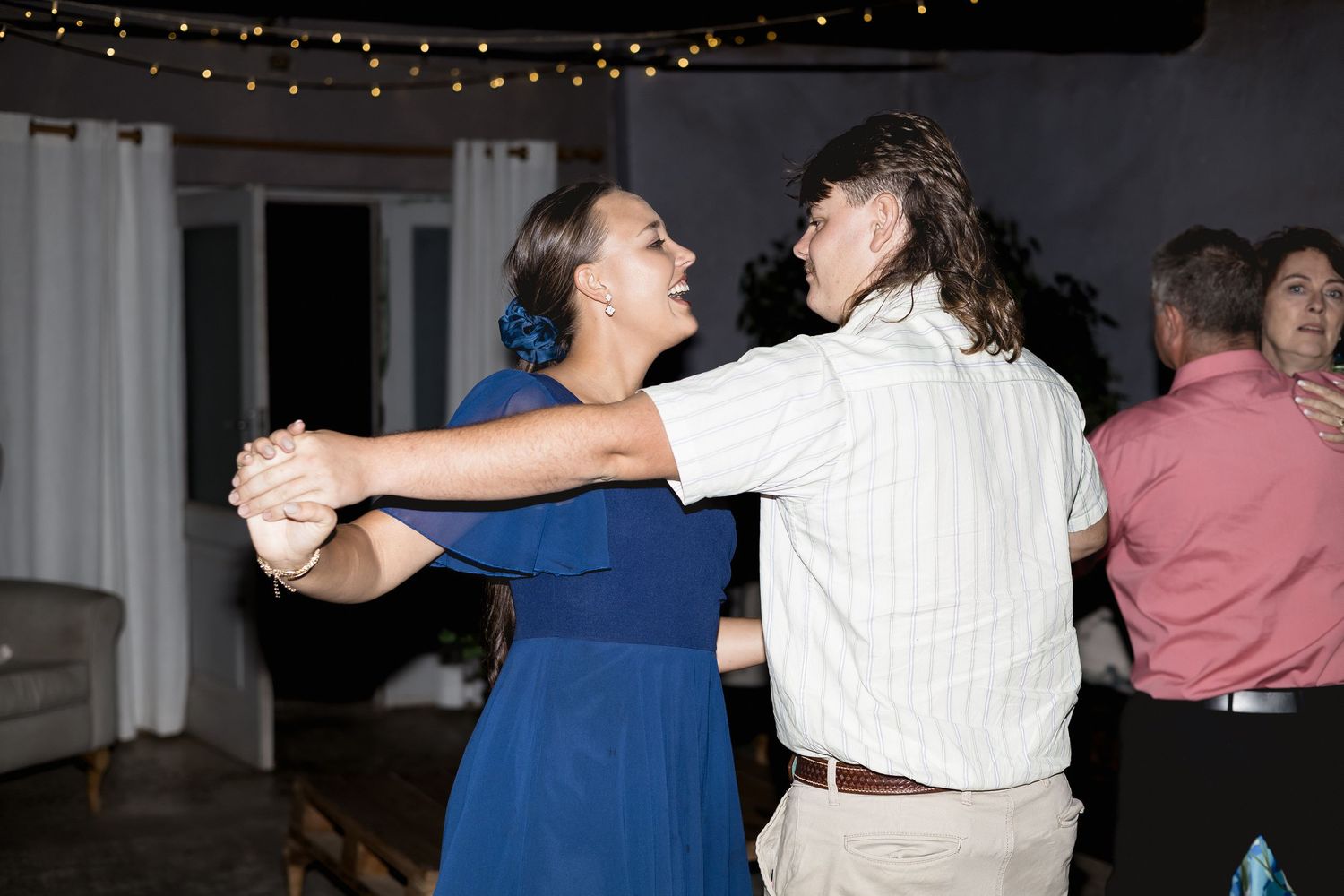 Couple dancing together at a wedding reception, woman in blue dress, man in white shirt, string lights above.