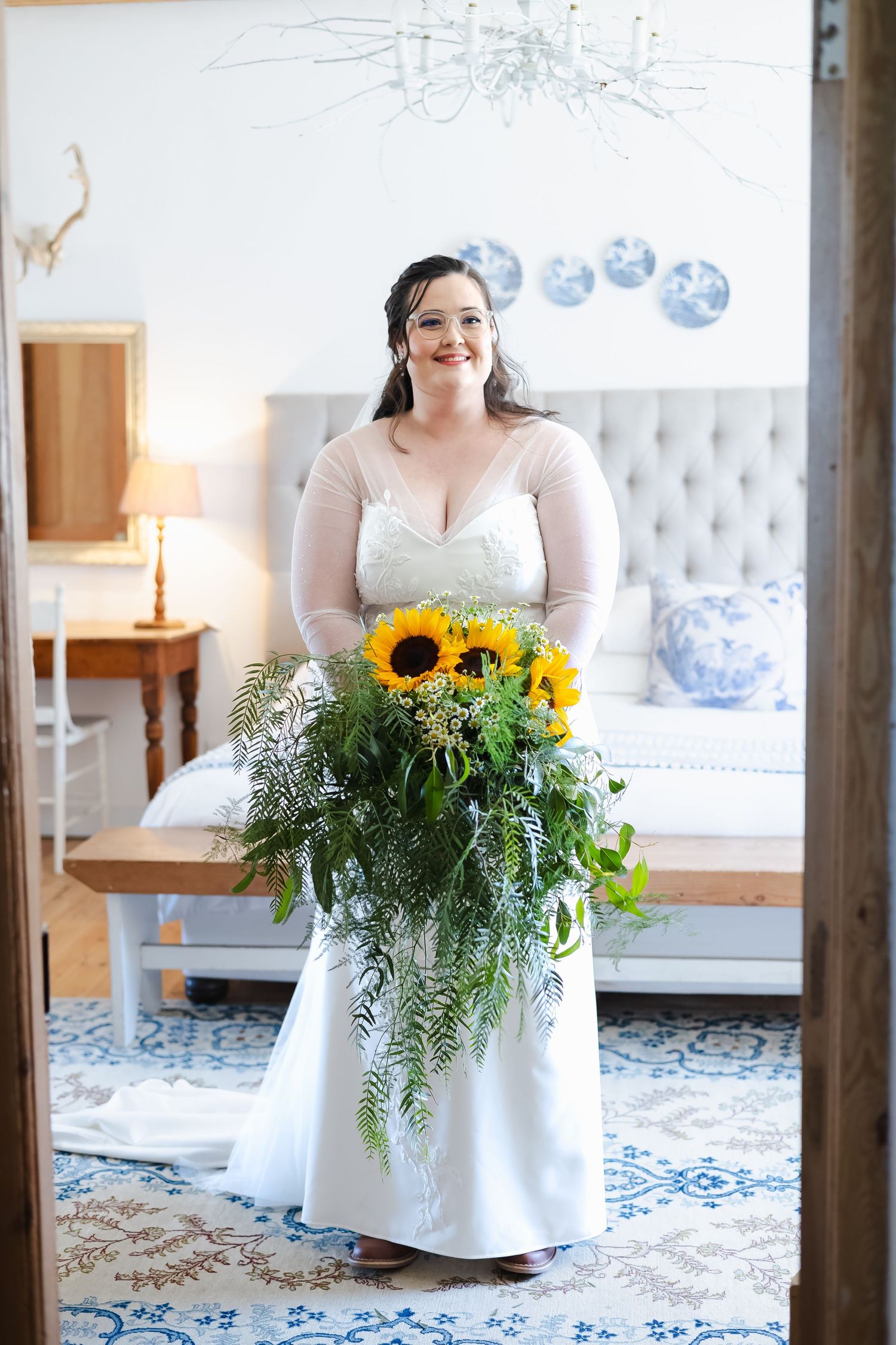Bride in white gown holds large sunflower and fern bouquet, standing in elegant bedroom doorway.