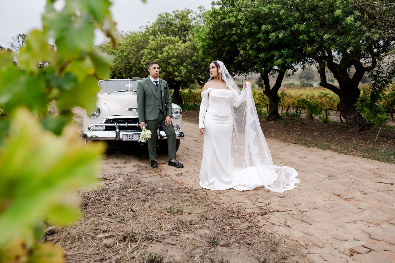 Bride in white gown and veil stands near groom beside vintage car in vineyard setting.