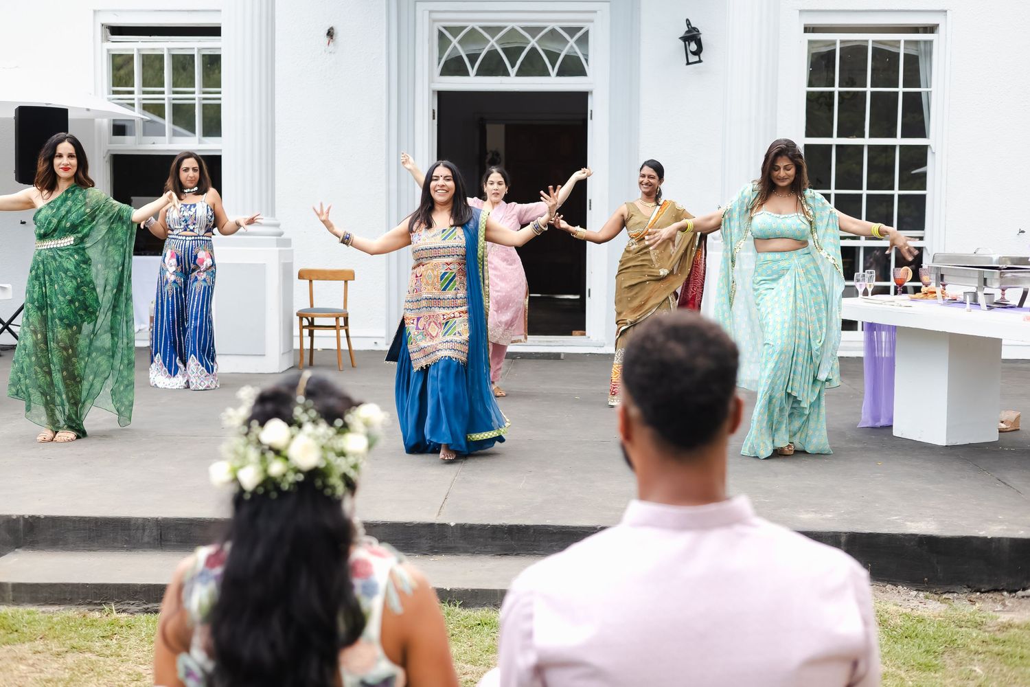 Wedding party members in mint green and blue dresses dance together on a lawn outside a white colonial building.
