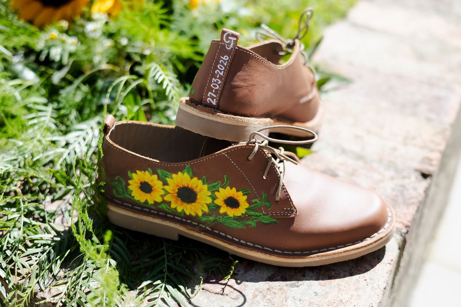 Brown leather lace-up shoes with hand-painted sunflower embroidery design, displayed outdoors among green foliage.