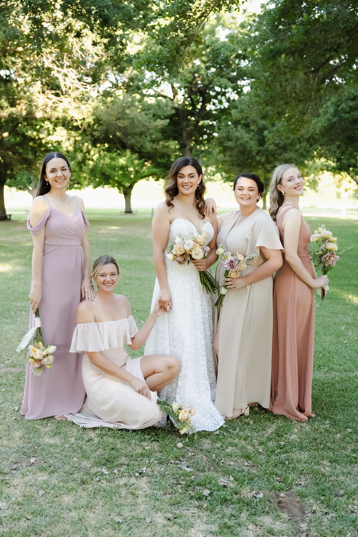 Bridesmaids in blush pink and neutral dresses gather outdoors for wedding photos on a sunny summer day.