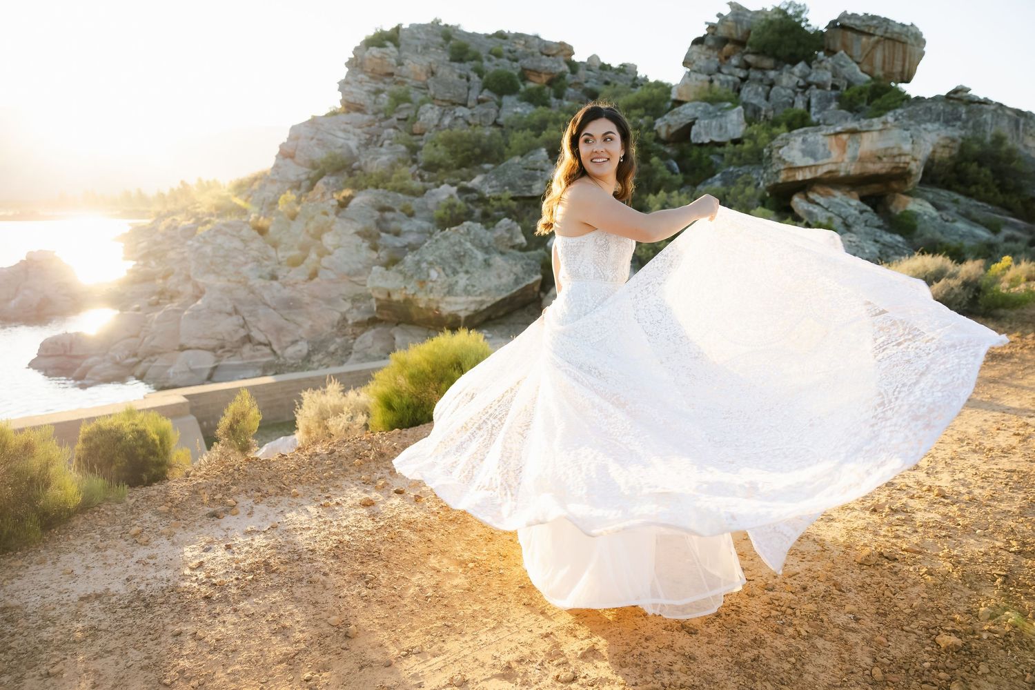 Bride twirls her flowing white dress against golden sunset and rocky coastal backdrop.