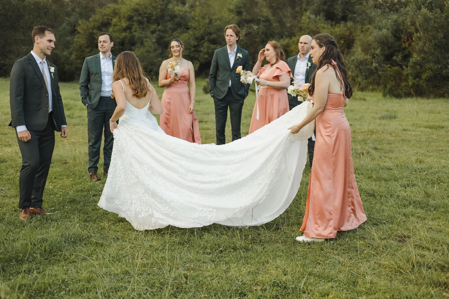 Wedding party members in coral dresses and suits pose together on a grassy field while holding a flowing white veil.
