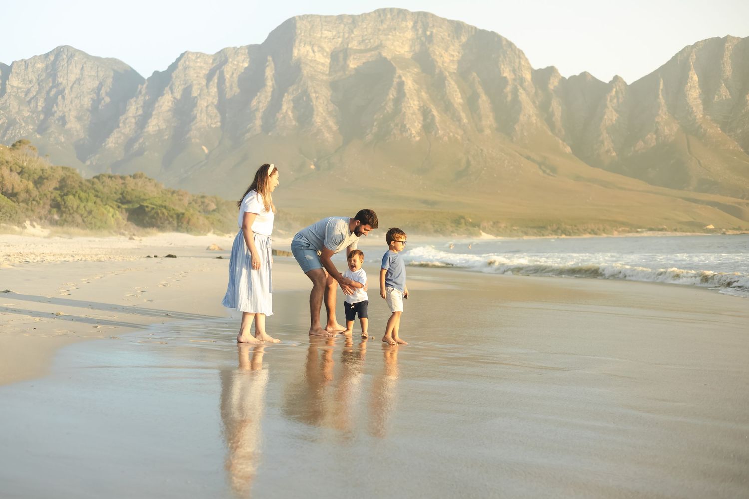 Family of four enjoying a sunny beach day with stunning mountain backdrop, children playing at the water's edge.