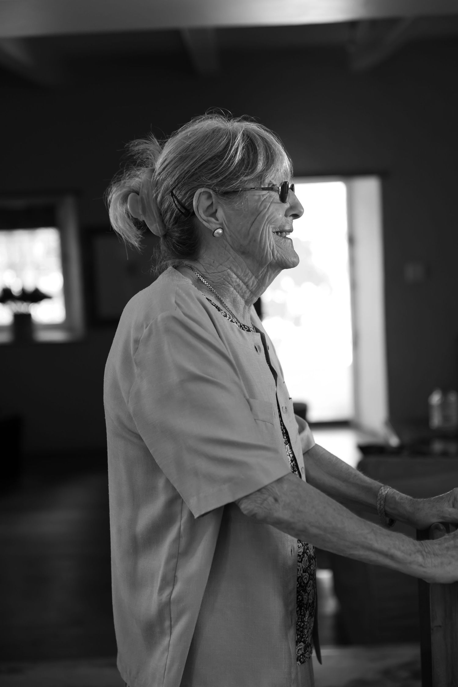 A black and white candid portrait of an elderly woman in glasses smiling while looking to the side.