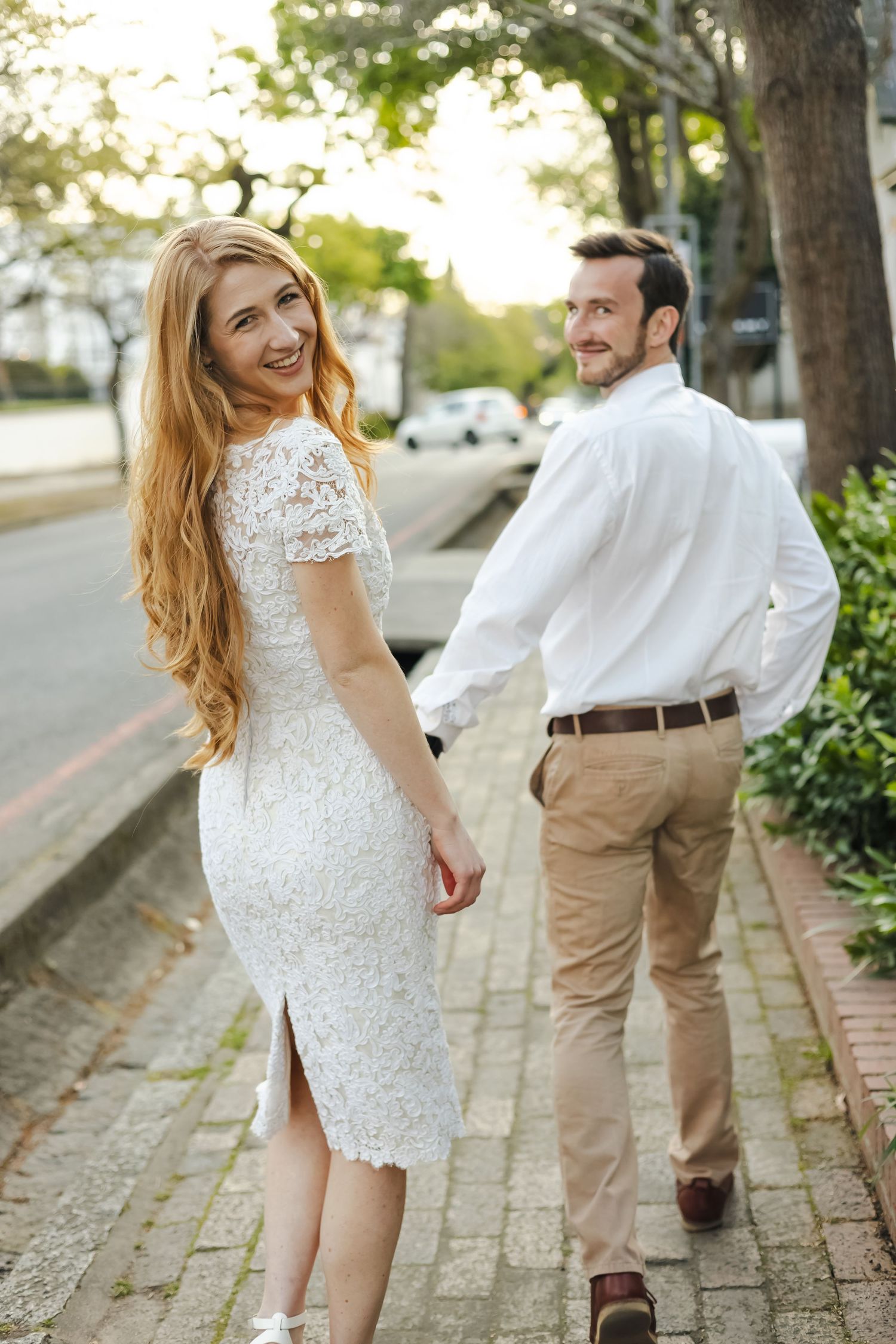 A couple walks together on a brick path, the bride in a white lace dress and groom in khaki pants and white shirt.