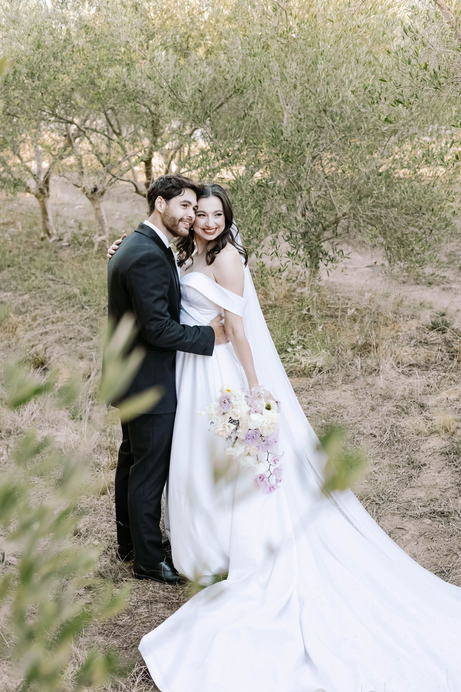 A couple in wedding attire poses together on natural terrain with desert vegetation.