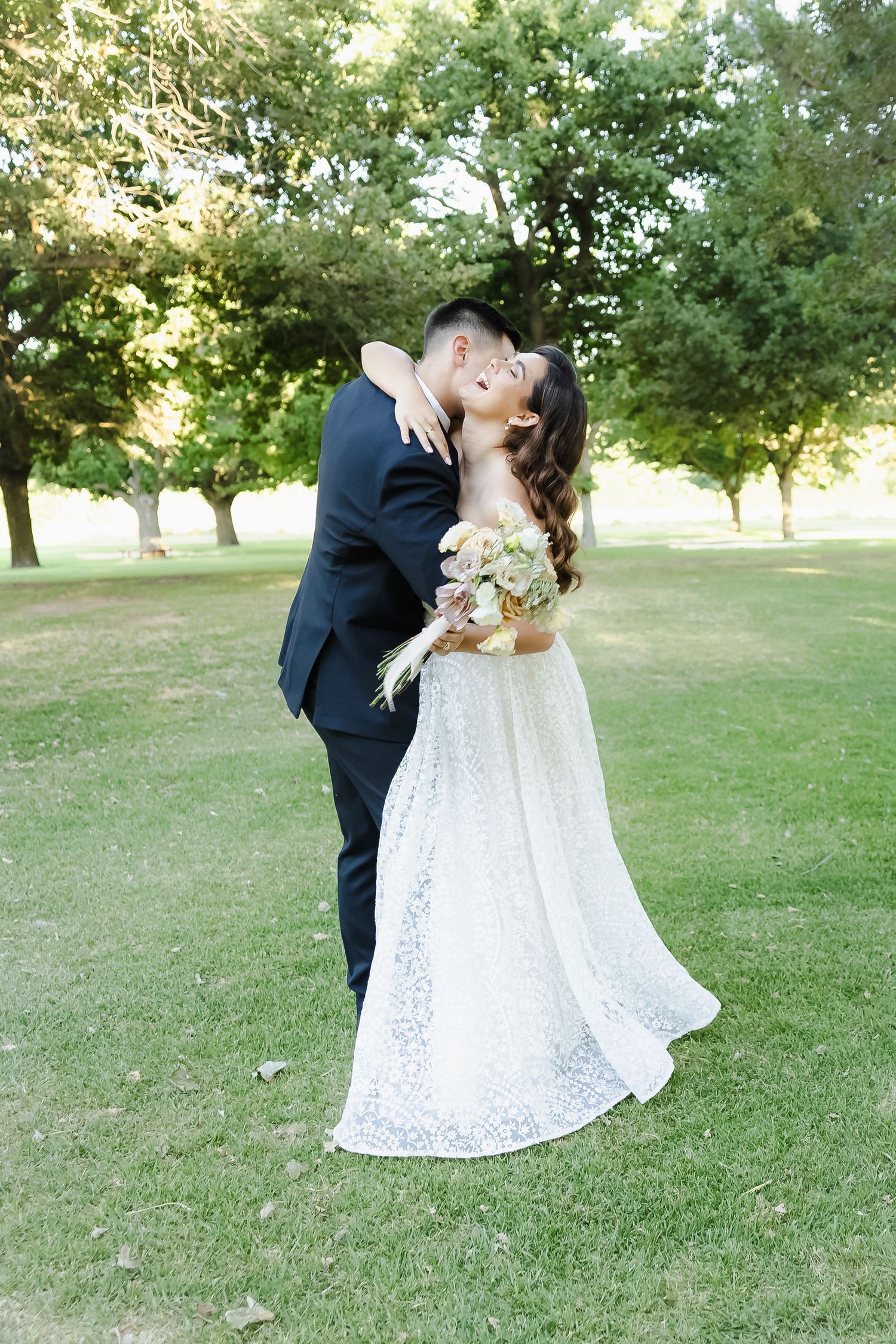 A romantic wedding couple embraces on a grassy lawn surrounded by trees during a sunny outdoor celebration.