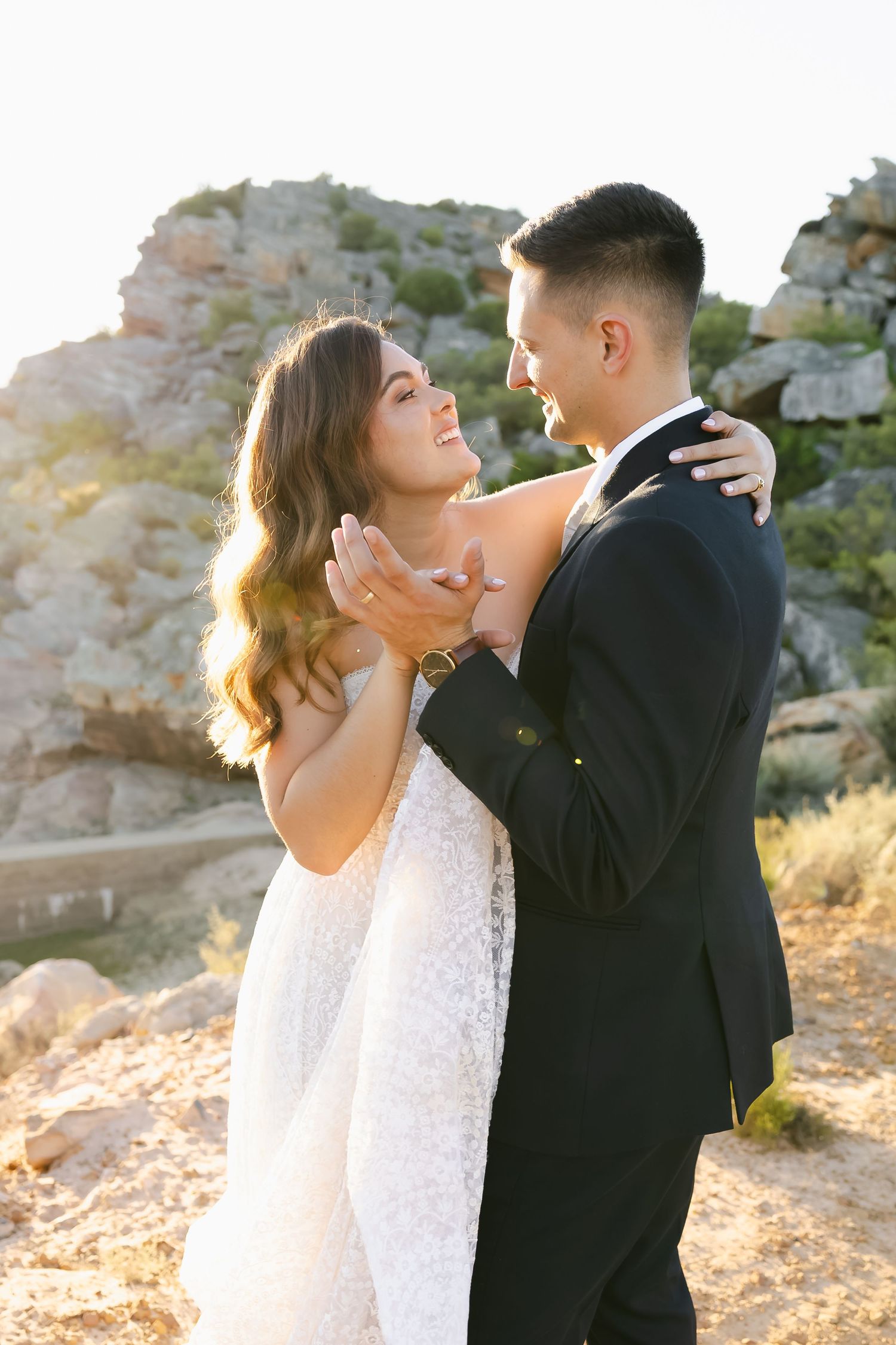 A romantic wedding couple embraces against a golden sunset backdrop in a desert setting.