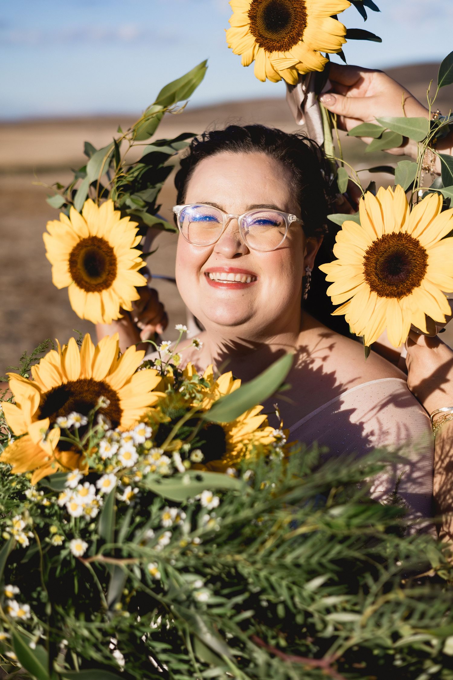 Smiling woman with glasses surrounded by vibrant sunflowers and greenery outdoors on a sunny day.