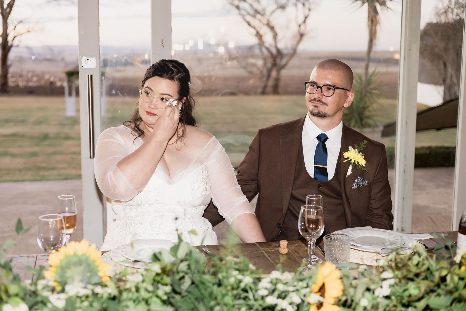 Bride wiping tears while groom sits beside her at wedding reception table decorated with sunflowers.
