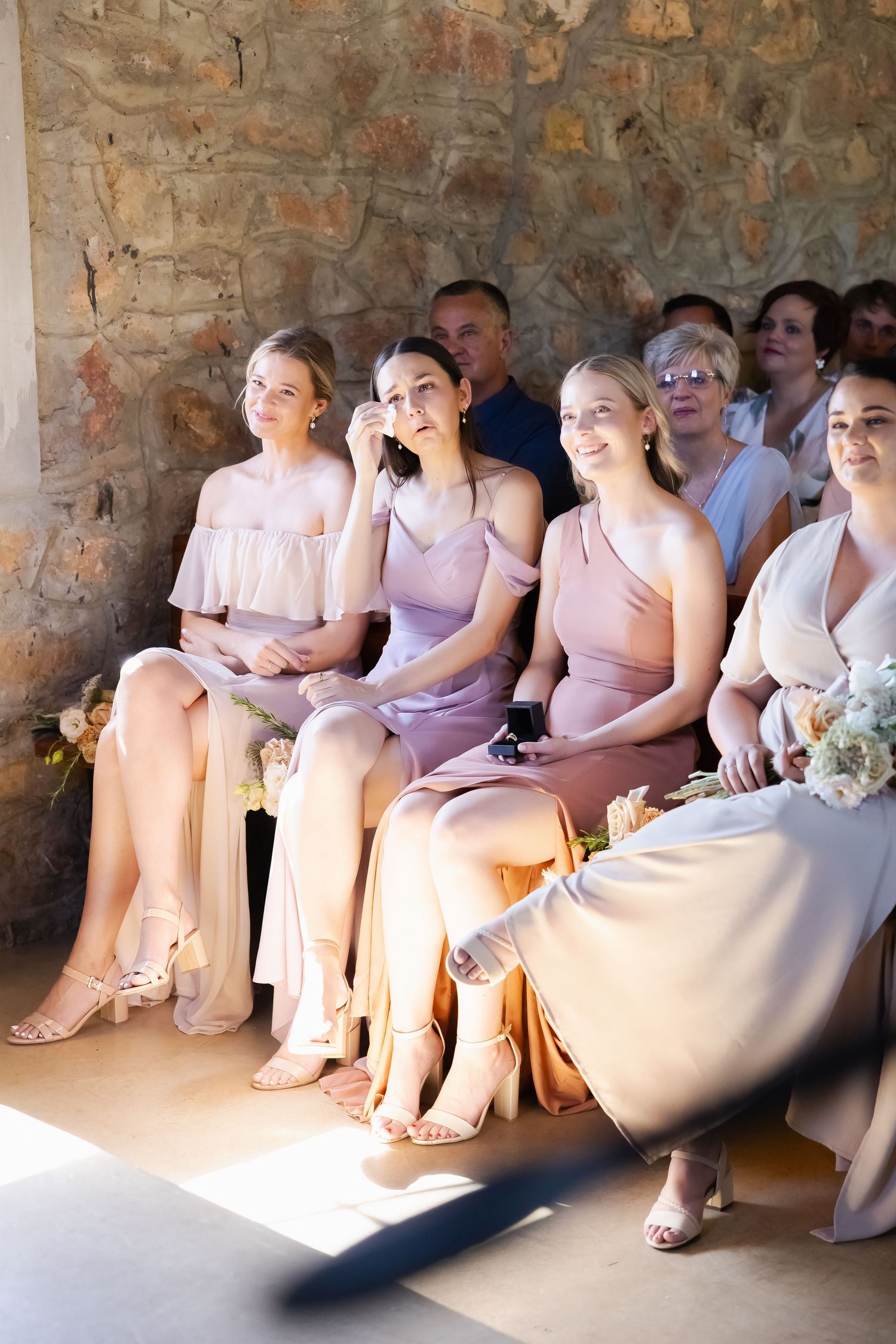 Wedding party members in soft pink and lavender dresses sit together on stone bench in sunlit corner.