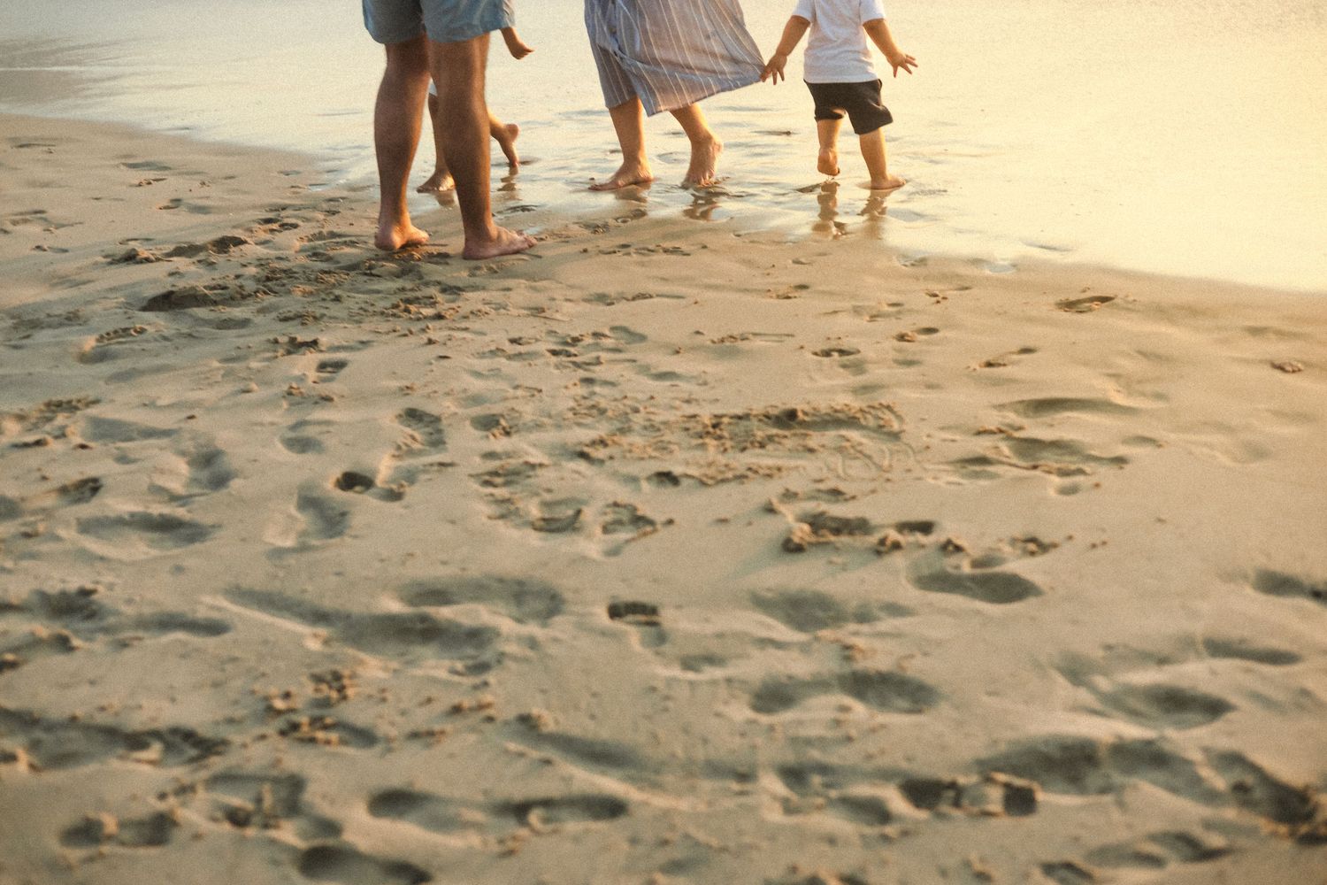 Family walking barefoot along a sandy beach shore leaving footprints in the sand.