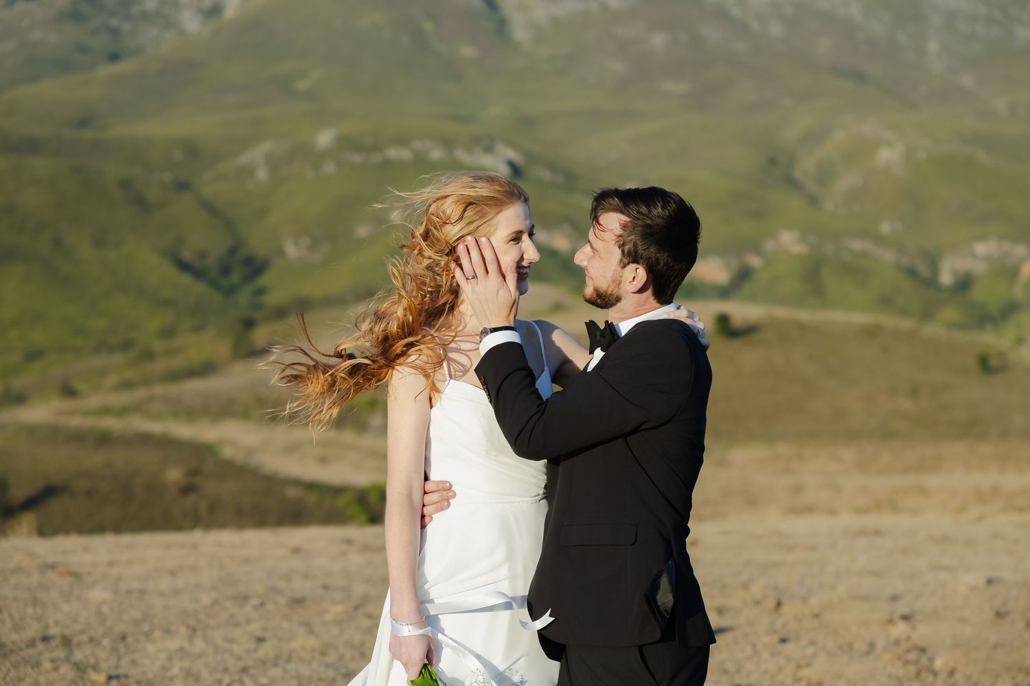 A couple in wedding attire shares an intimate moment against a backdrop of rolling green hills and mountains.