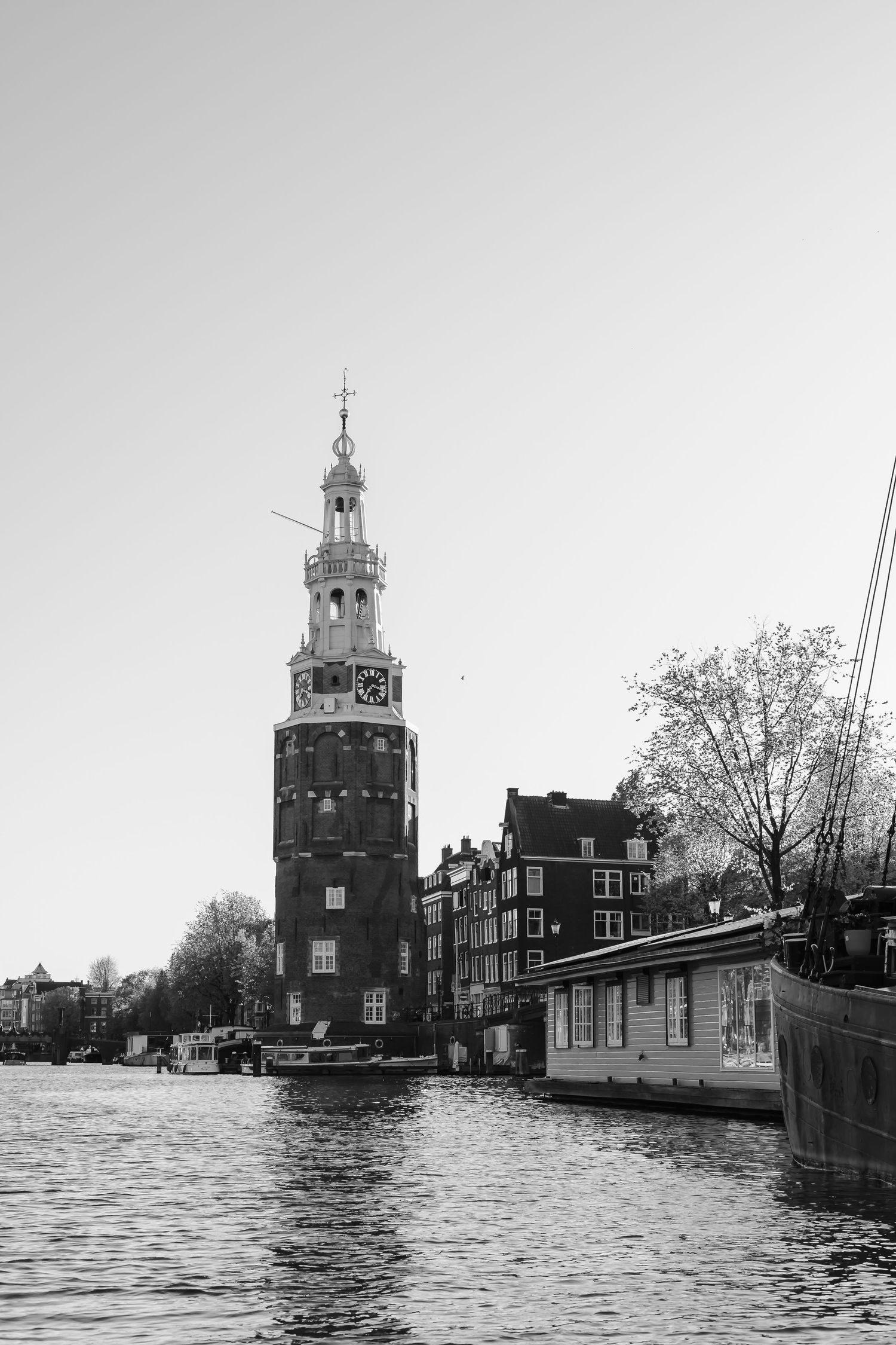 Black and white photo of Amsterdam canal with the historic Montelbaanstoren tower and moored houseboats on a clear day.