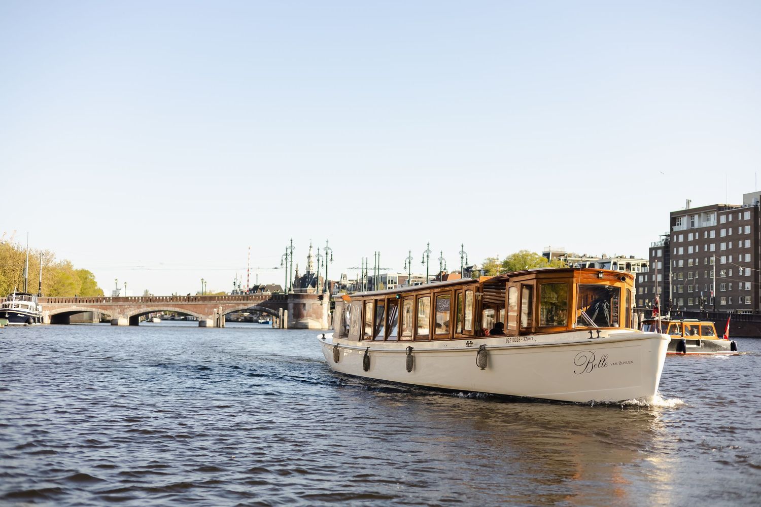A classic white canal boat cruising on the Amsterdam river with a bridge and city buildings in the background.