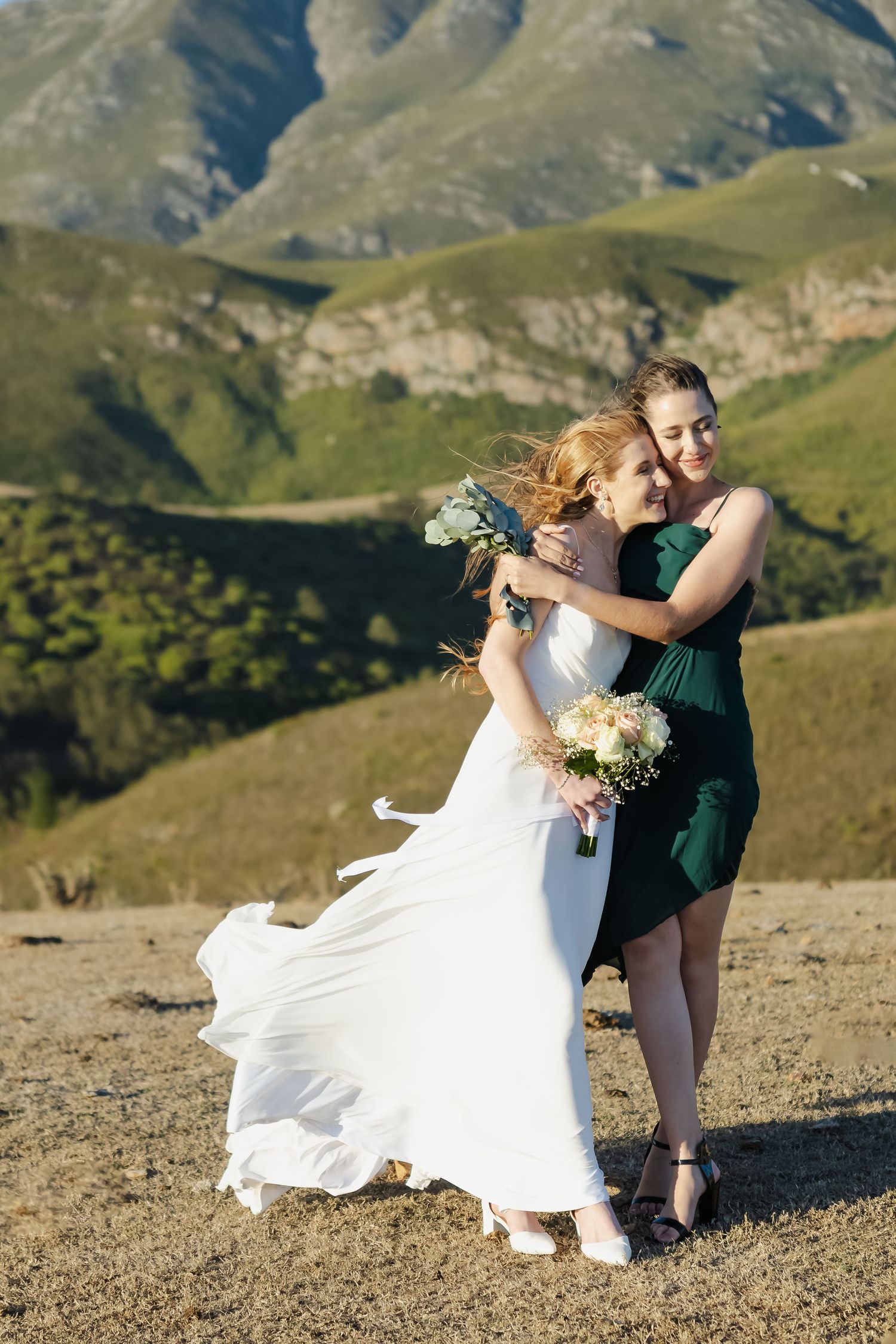 Two people embrace in formal attire against a dramatic mountain backdrop as a white dress flows in the wind.