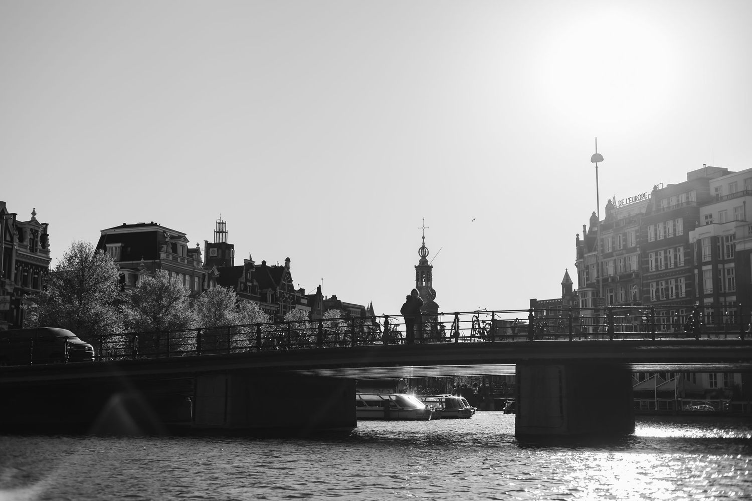 Black and white photo of a canal bridge in Amsterdam with a boat passing underneath, historic buildings and church tower in background.