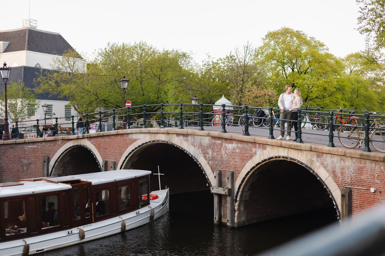 People walking on a historic brick arched bridge over an Amsterdam canal with a boat below and lush green trees.