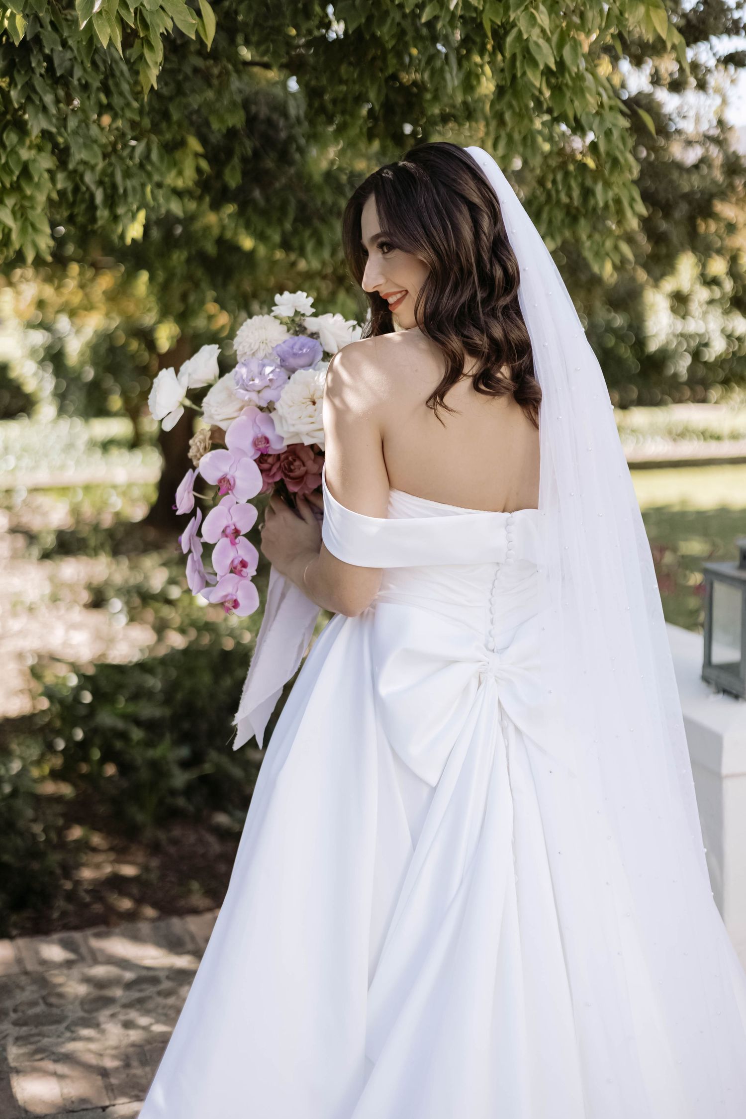 Close-up details of a white wedding dress with bow accents and orchid bouquet from behind.