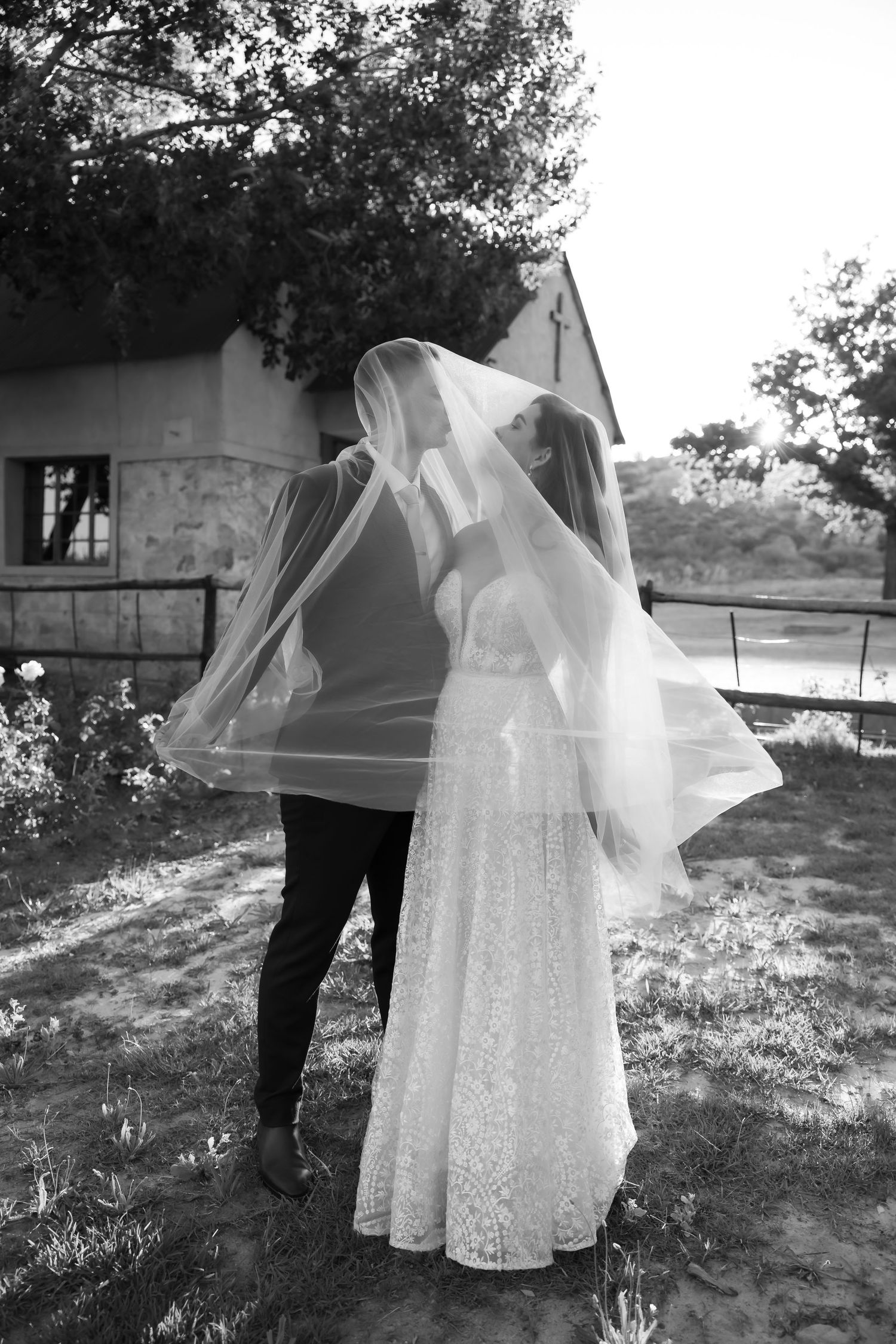 A black and white wedding photo showing a couple embracing outdoors with the bride wearing a flowing veil and lace dress.