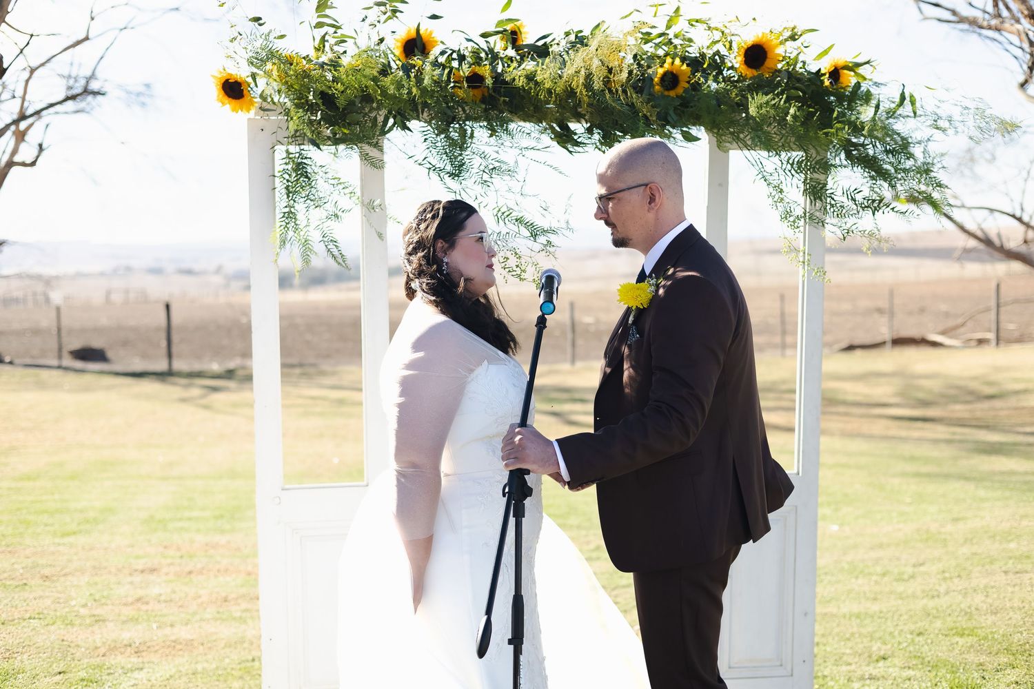 Couple exchanging vows under sunflower-adorned white arch at outdoor wedding ceremony on green lawn.