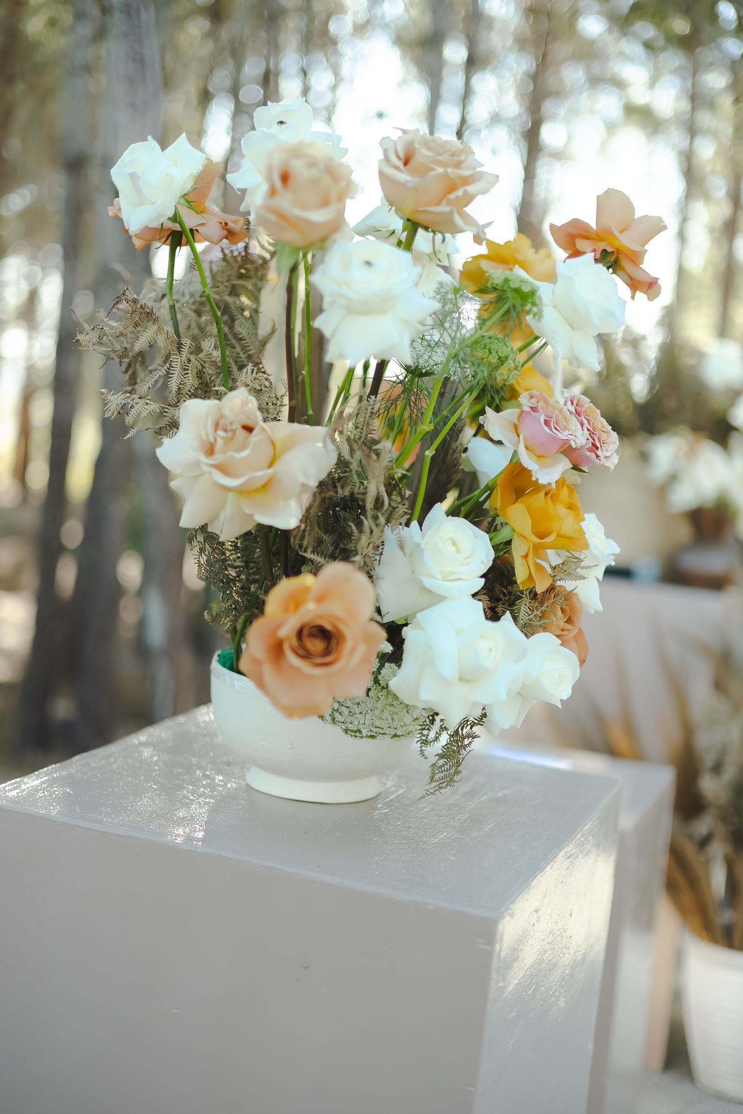Elegant floral arrangement of white and peach roses with greenery displayed on white pedestals in a sunlit outdoor setting.