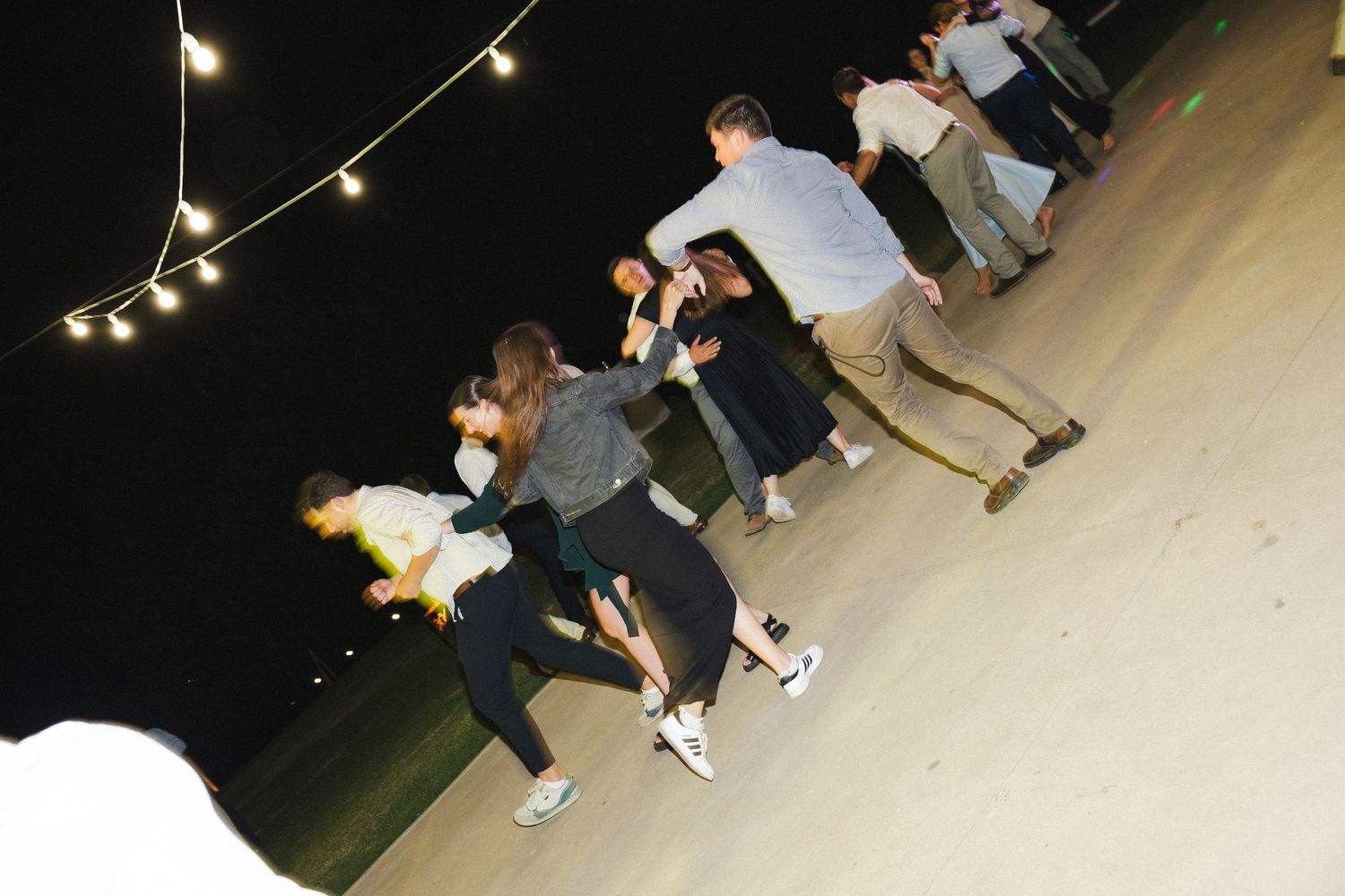 Skateboarders practicing tricks at night under string lights in an outdoor skate park.