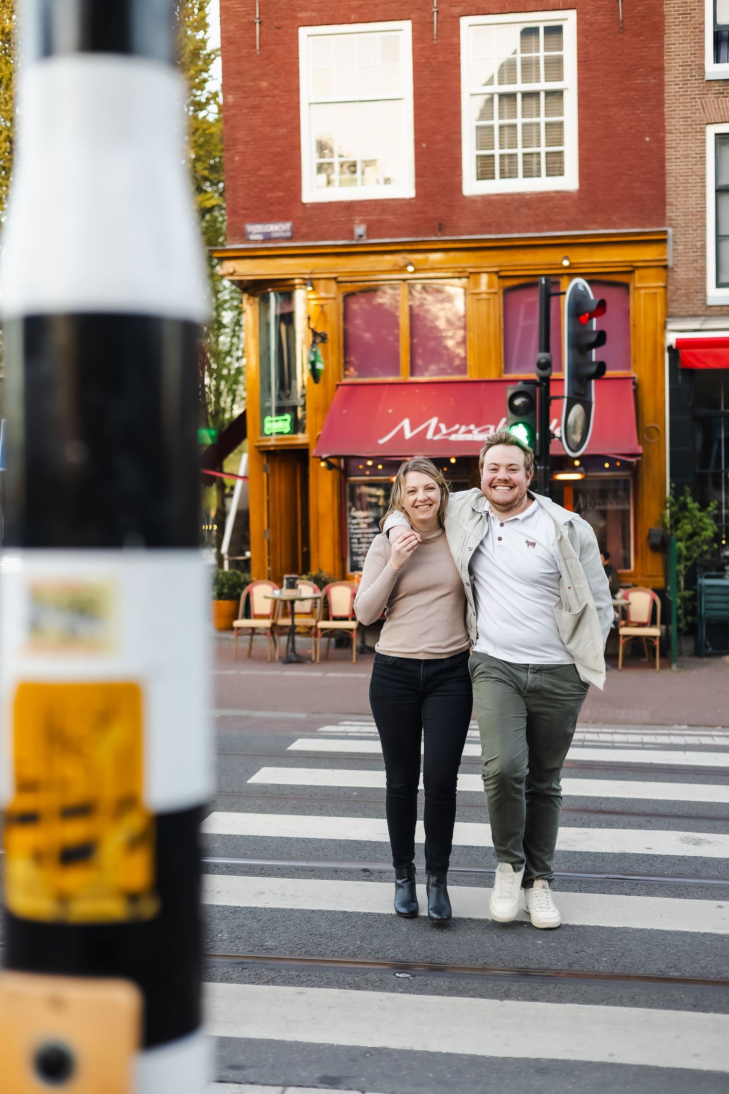 A smiling couple walks across a zebra crossing in Amsterdam with a colorful restaurant facade behind them.