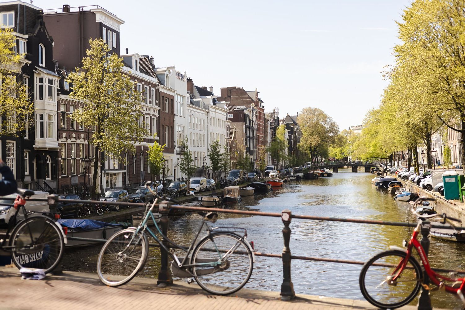 Bicycles parked on a bridge over an Amsterdam canal lined with historic buildings and boats on a sunny day.