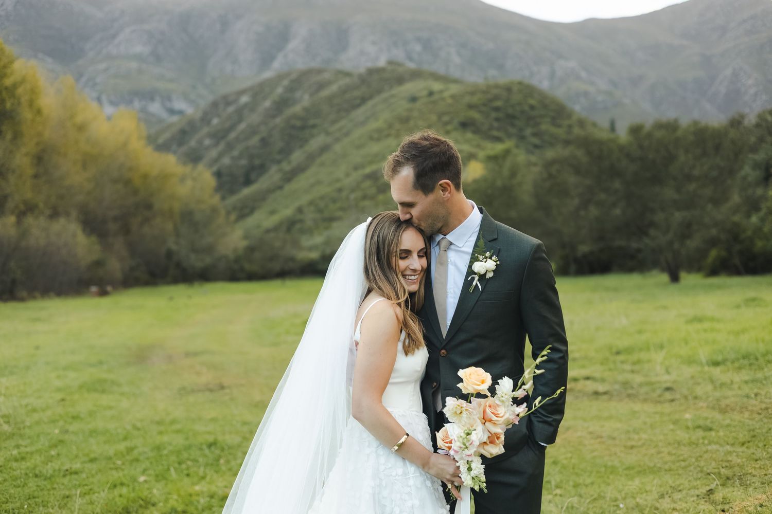 A romantic wedding portrait shows a couple embracing in a lush green mountain meadow with white florals and a flowing veil.